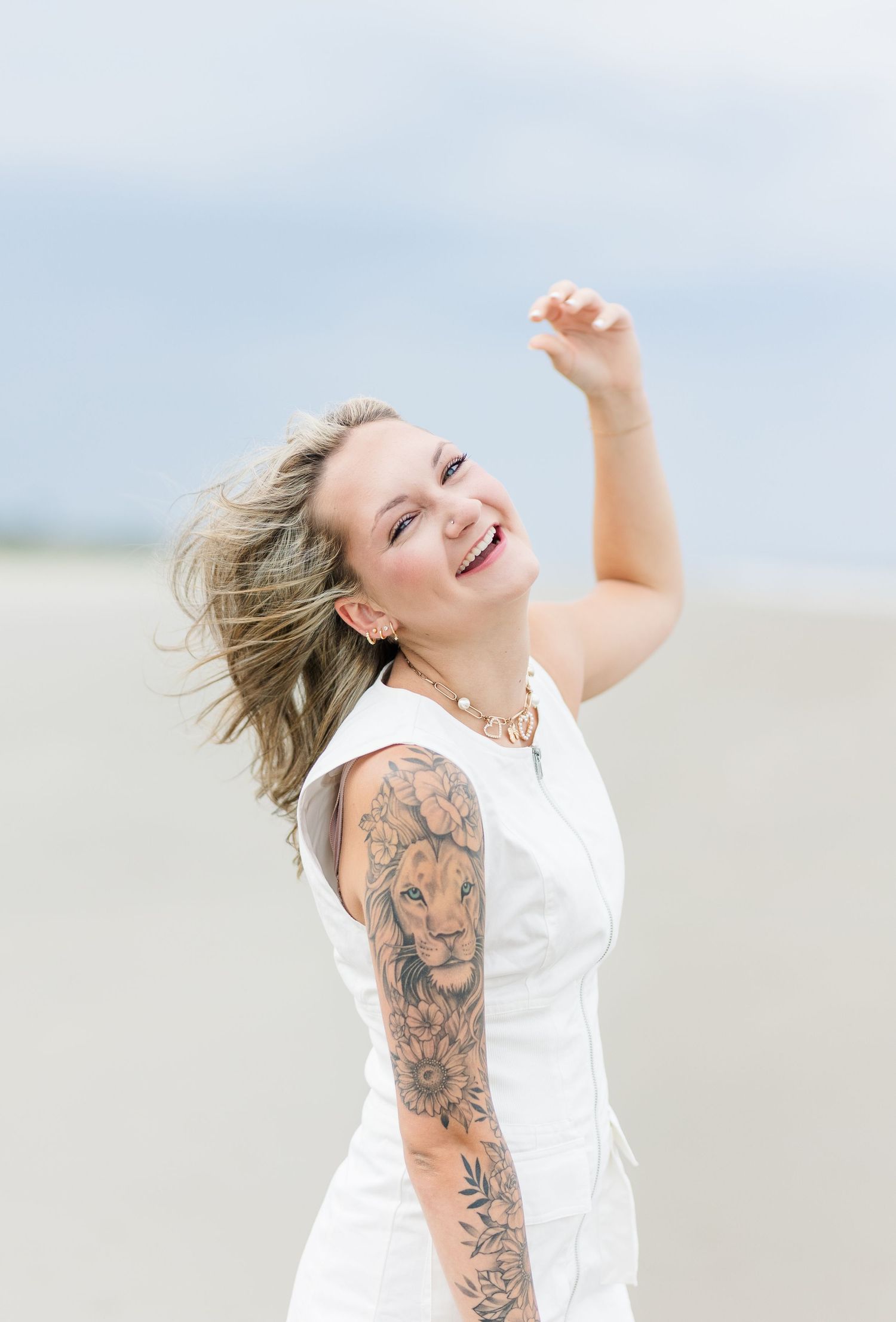 A person in a white dress dances joyfully on a beach with tattoos visible on their arm.