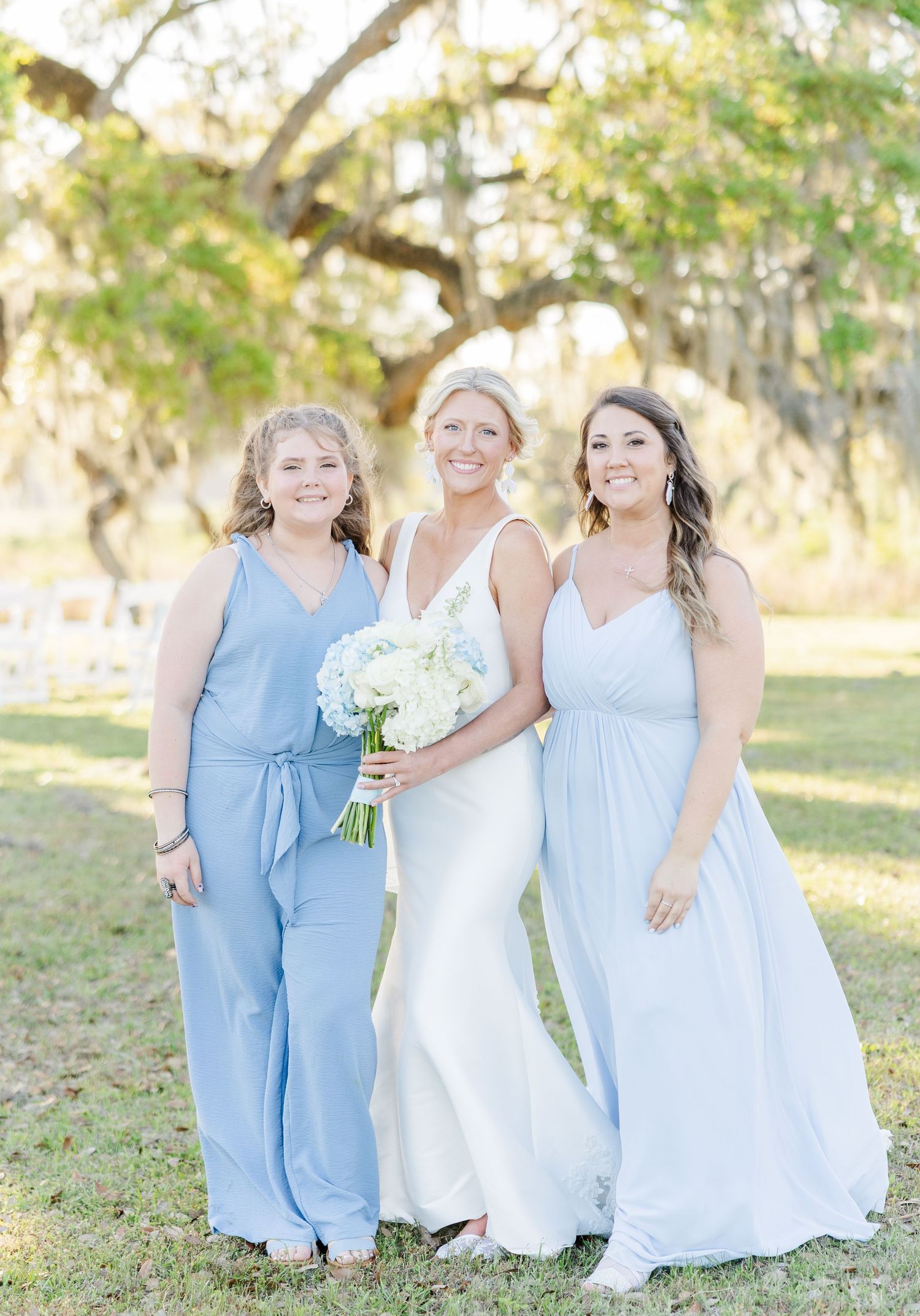 Bride in white gown stands with two bridesmaids in blue attire under Spanish moss oak trees.