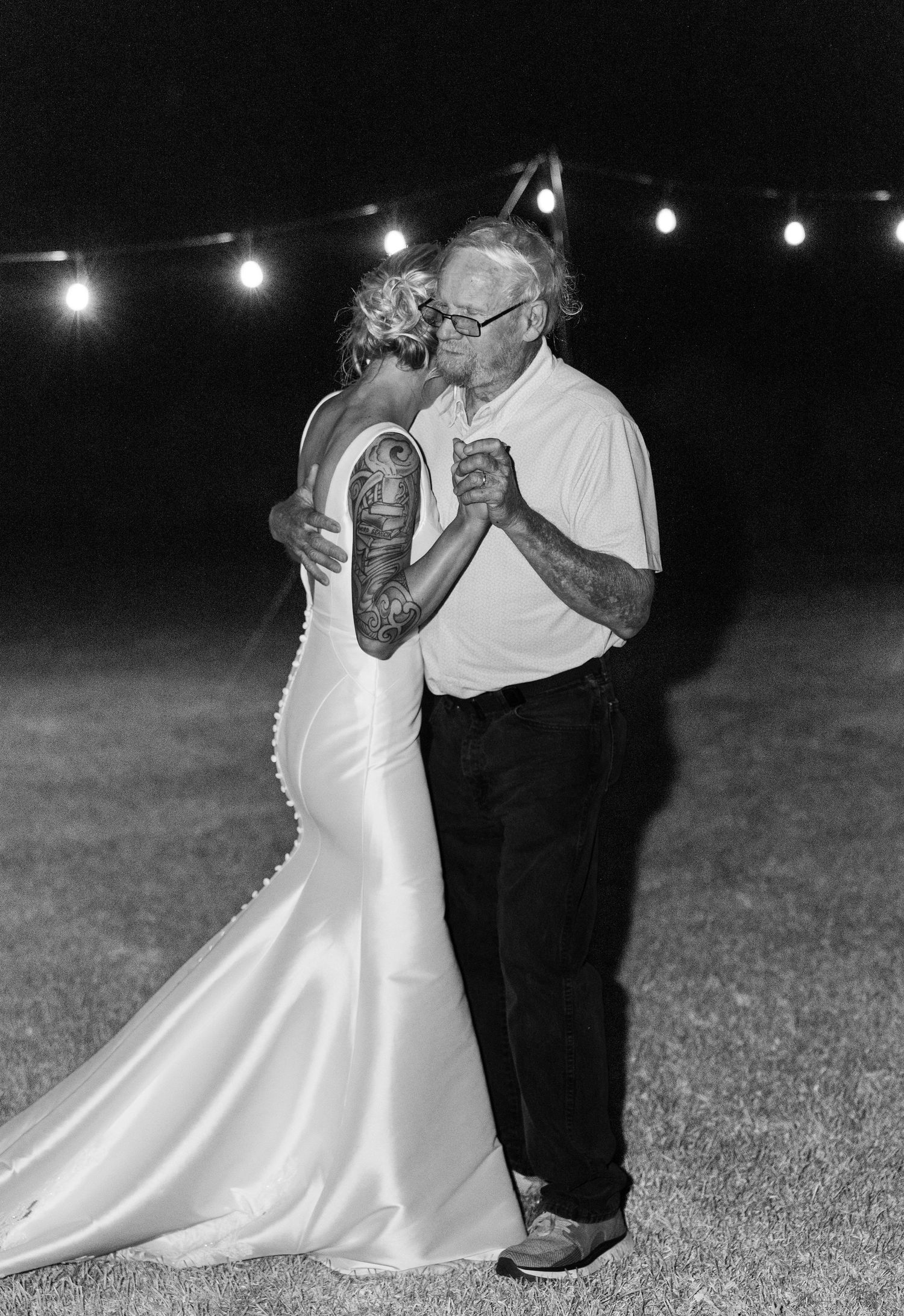 Black and white photo of bride in fitted gown dancing with elderly man under string lights at night.