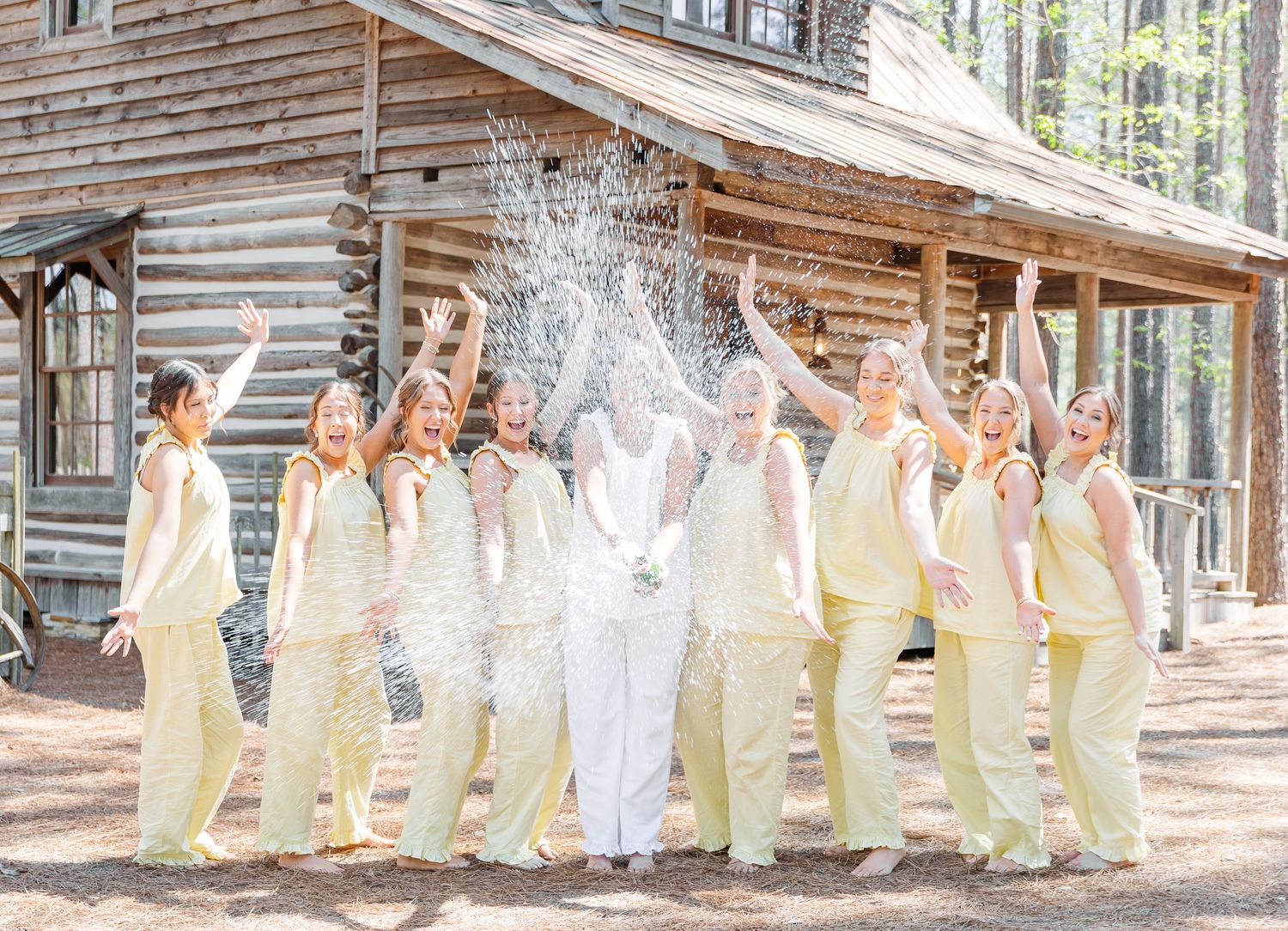 Bride and bridesmaids in yellow celebrate with a champagne spray outside a rustic log cabin venue.