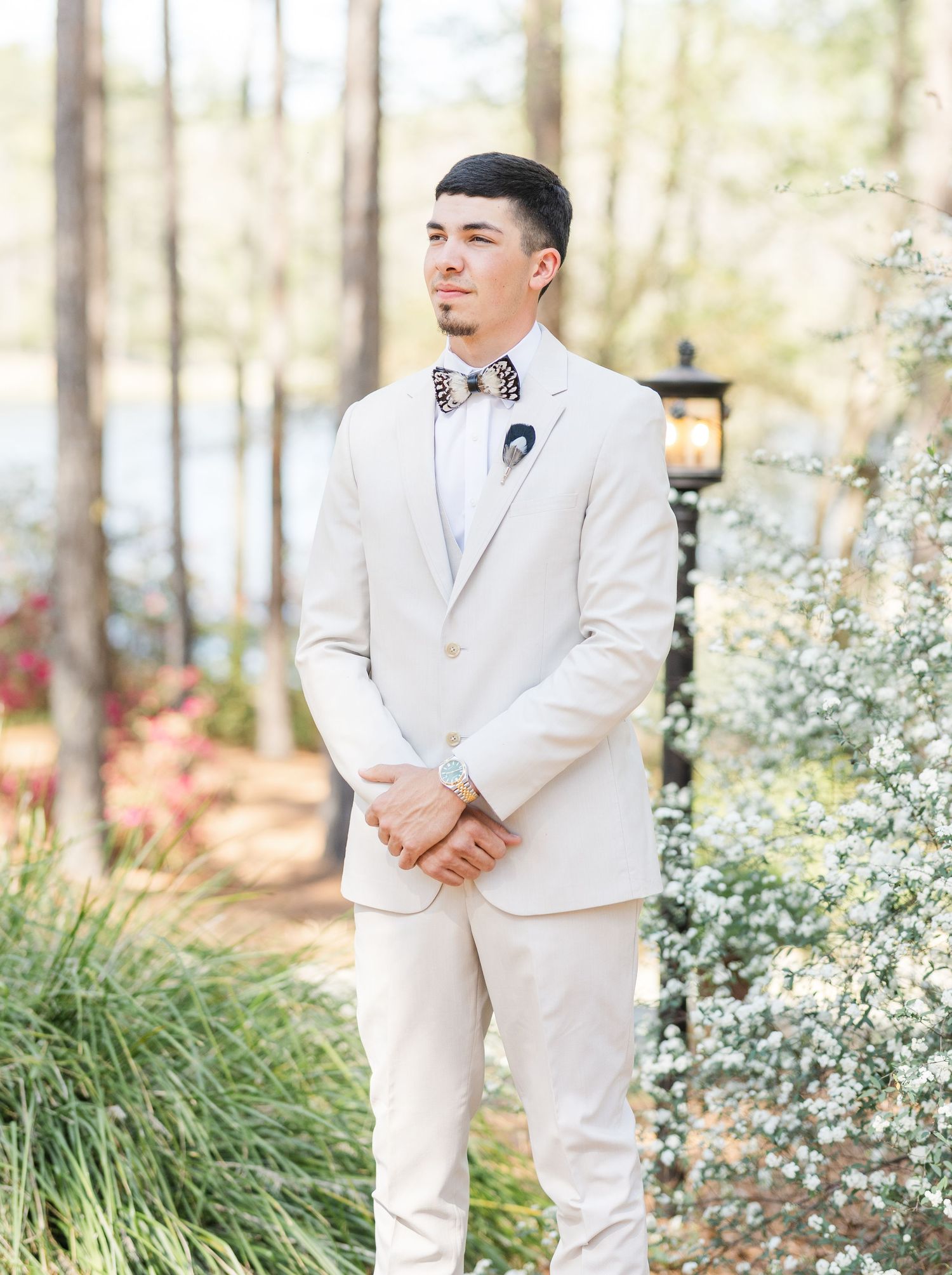 Groom in cream suit with patterned bow tie standing in garden before wedding ceremony.