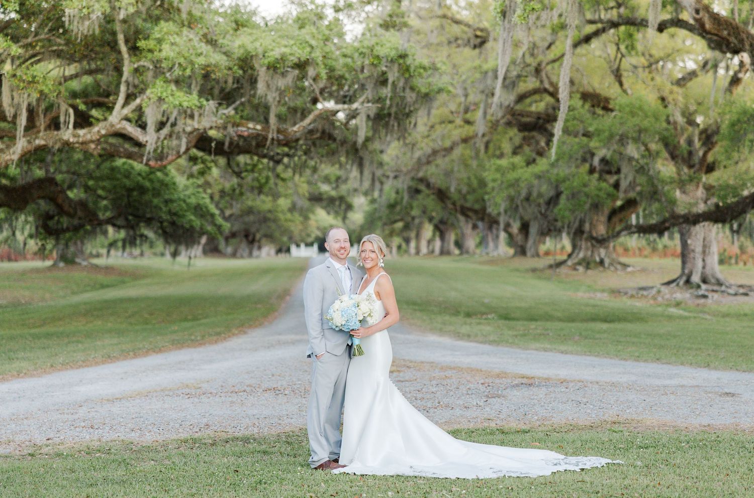 Bride and groom posing on a scenic tree-lined path with Spanish moss at outdoor wedding venue.