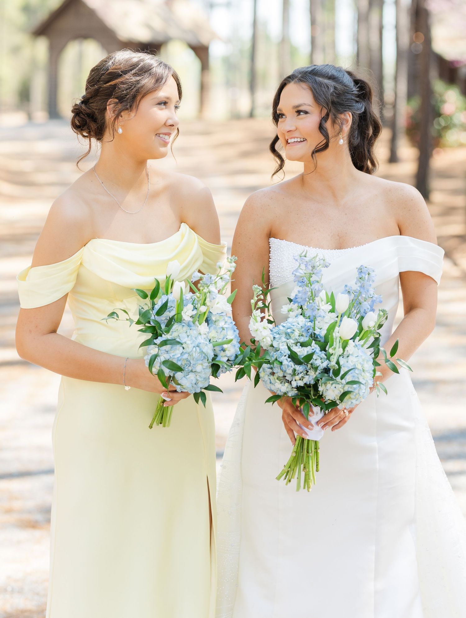 Bride and bridesmaid in soft yellow dress smile at each other holding blue flower bouquets.