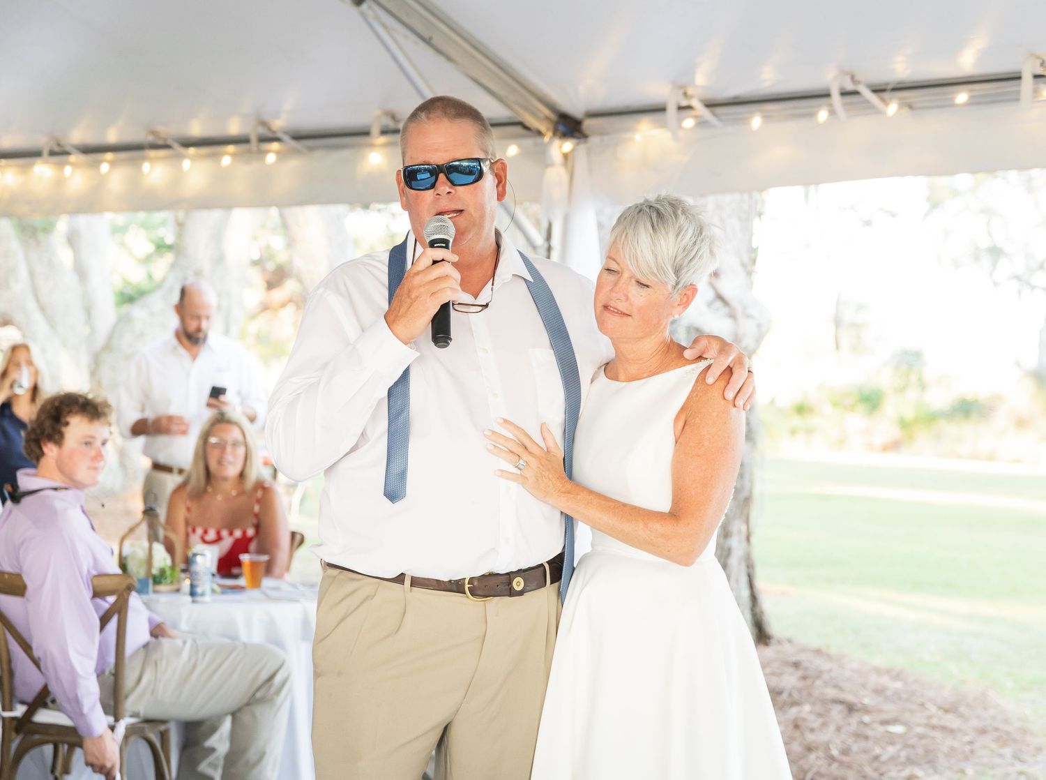 A couple shares a joyful moment under a white tent during a wedding reception as they speak into a microphone.