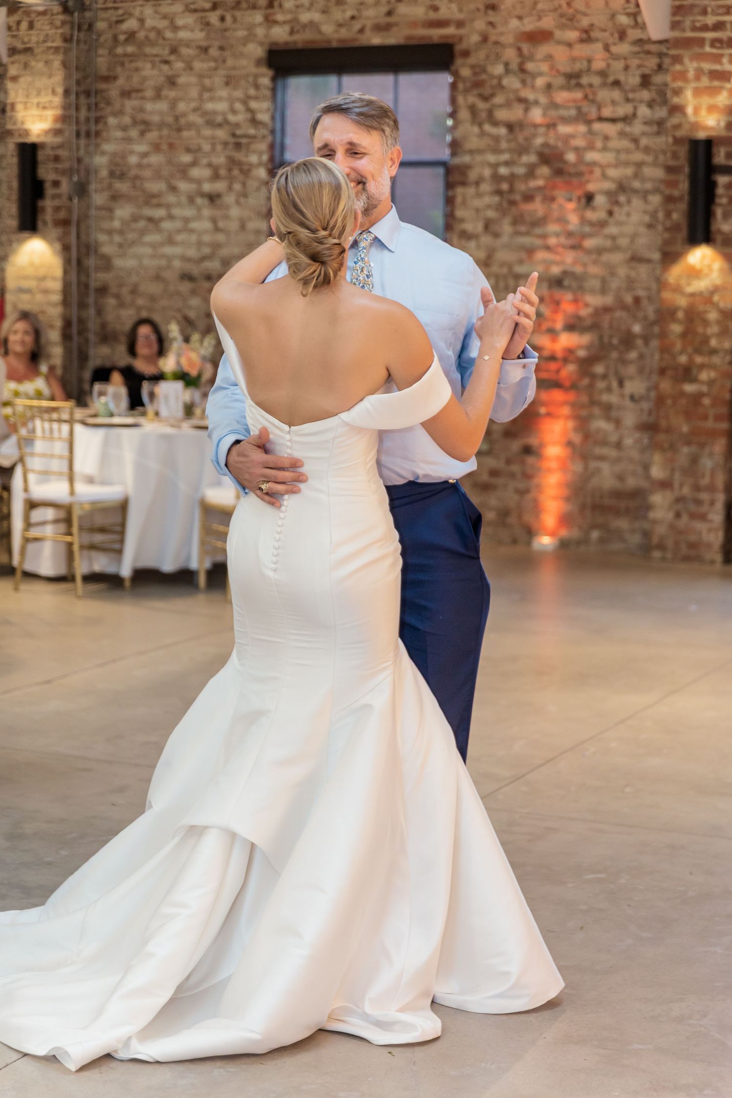 A couple shares their first dance at a rustic brick venue, with the bride wearing an elegant off-shoulder mermaid wedding gown.
