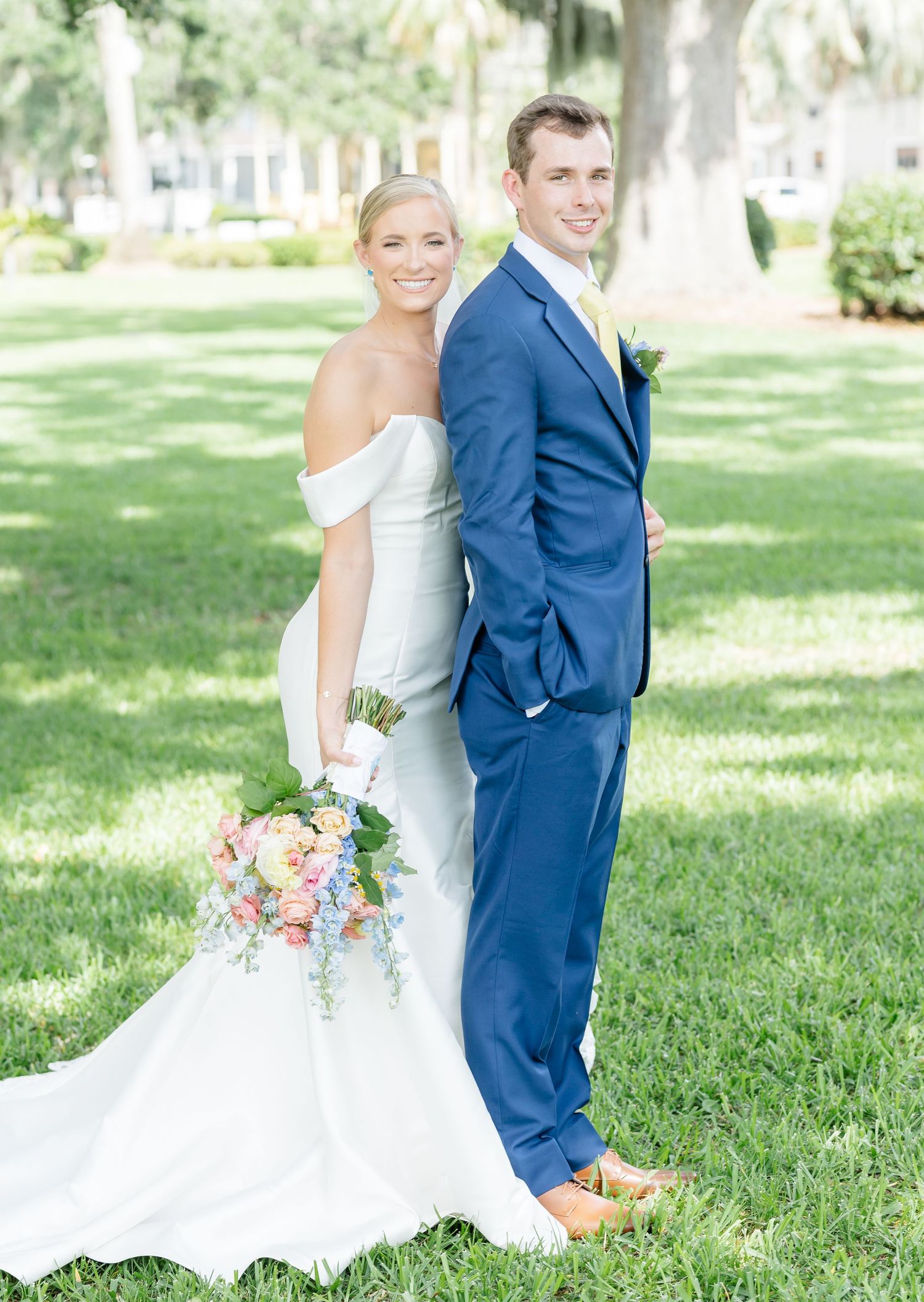 A bride in a white off-shoulder gown and groom in navy suit pose back-to-back on a sunny lawn with palm trees.