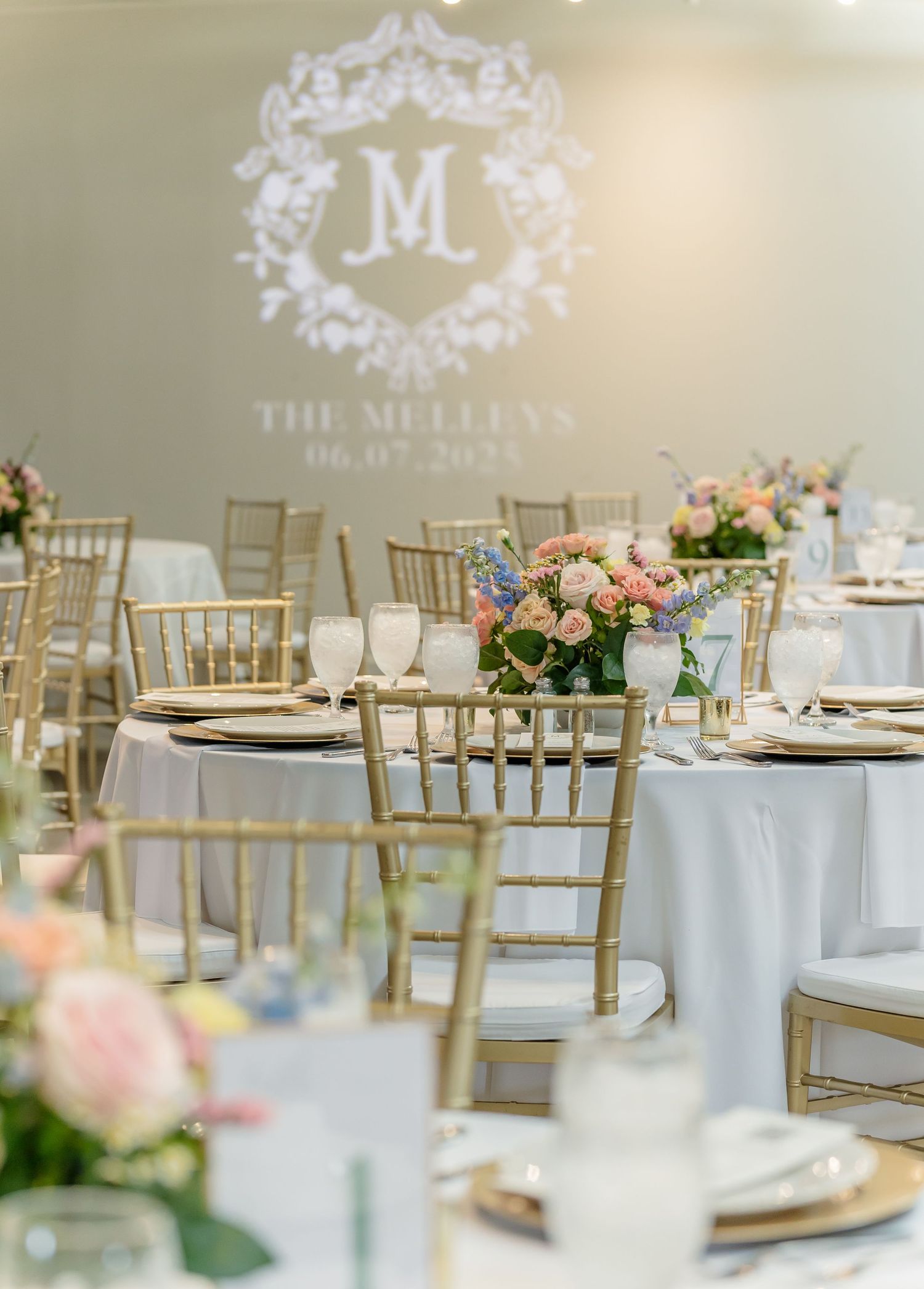 Elegant wedding reception setup with gold chiavari chairs, white tablecloths, and monogram lighting projection on the wall.