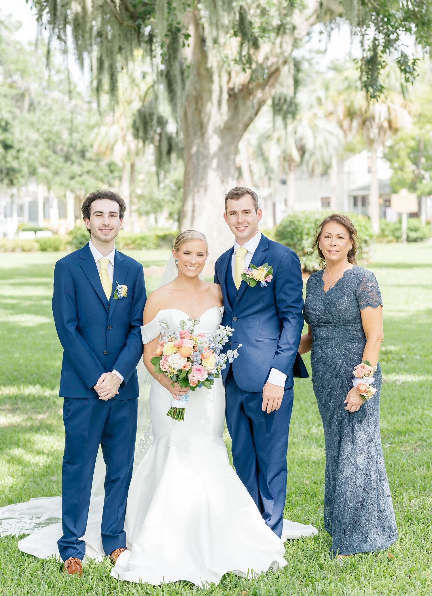 A wedding party in navy blue suits and formal attire poses together on a lush green lawn with Spanish moss trees.