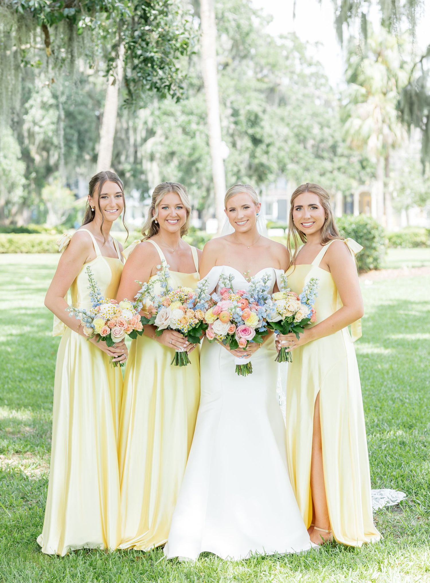 Wedding party in pastel yellow bridesmaid dresses poses with bouquets in a sunny garden setting.