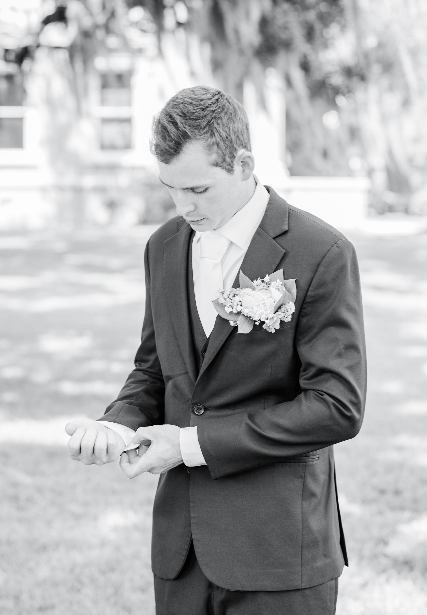 A groom in a dark suit with a boutonniere checks his watch while standing outdoors in a black and white photograph.