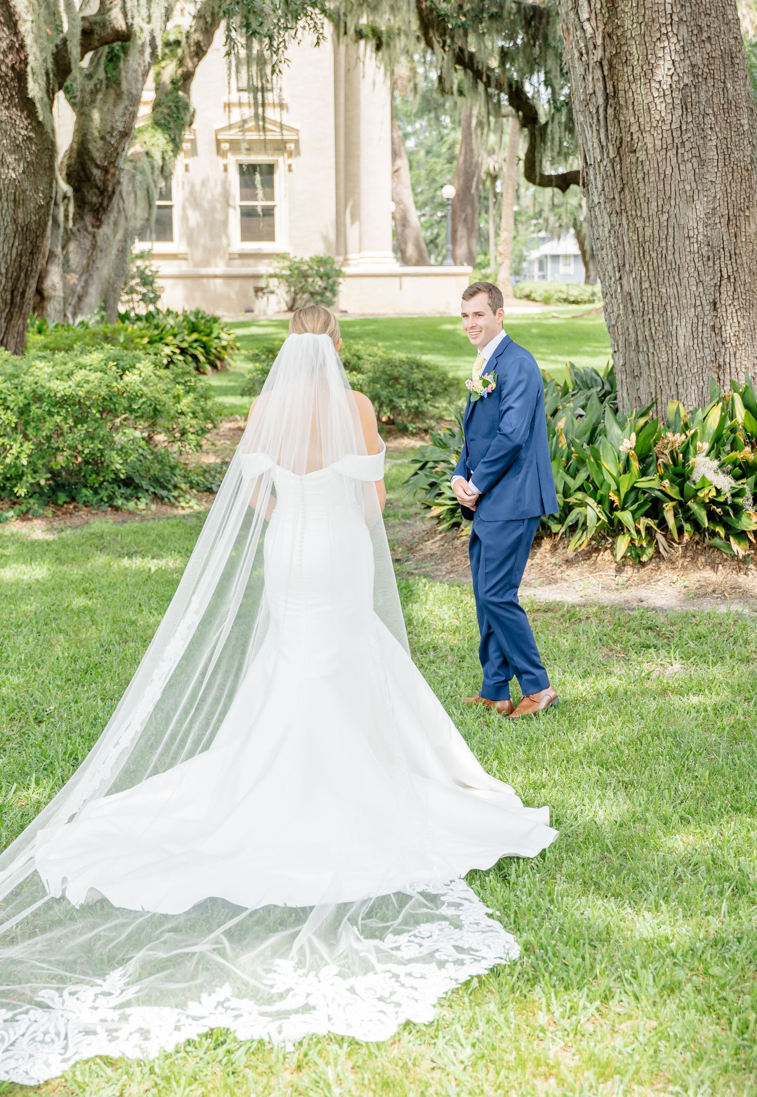 A wedding couple shares their first look moment under Spanish moss draped trees on a manicured lawn.