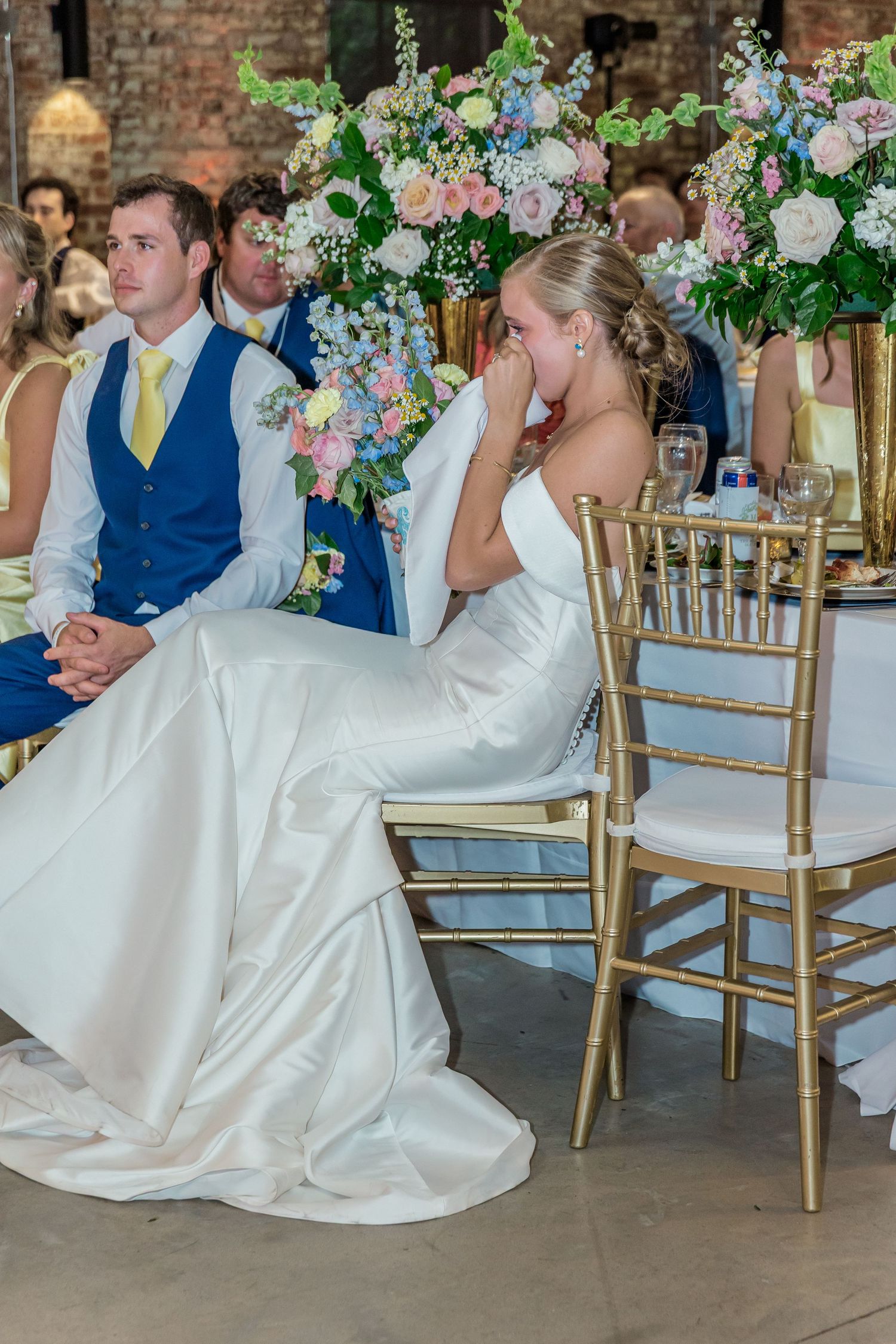 Elegant wedding reception scene with white floral arrangements and gold chairs in a rustic brick venue.