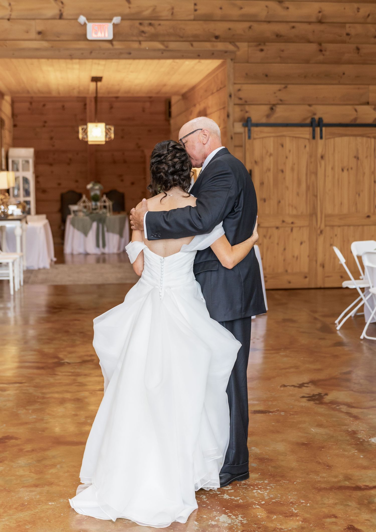 A couple shares a romantic dance in a rustic wooden reception hall with warm lighting and polished floors.