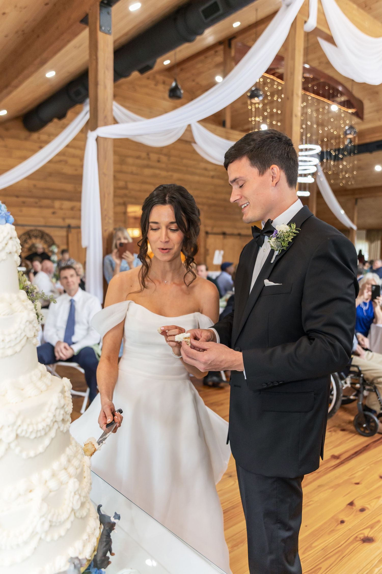 A couple cuts their elegant white wedding cake together in a rustic barn reception venue.