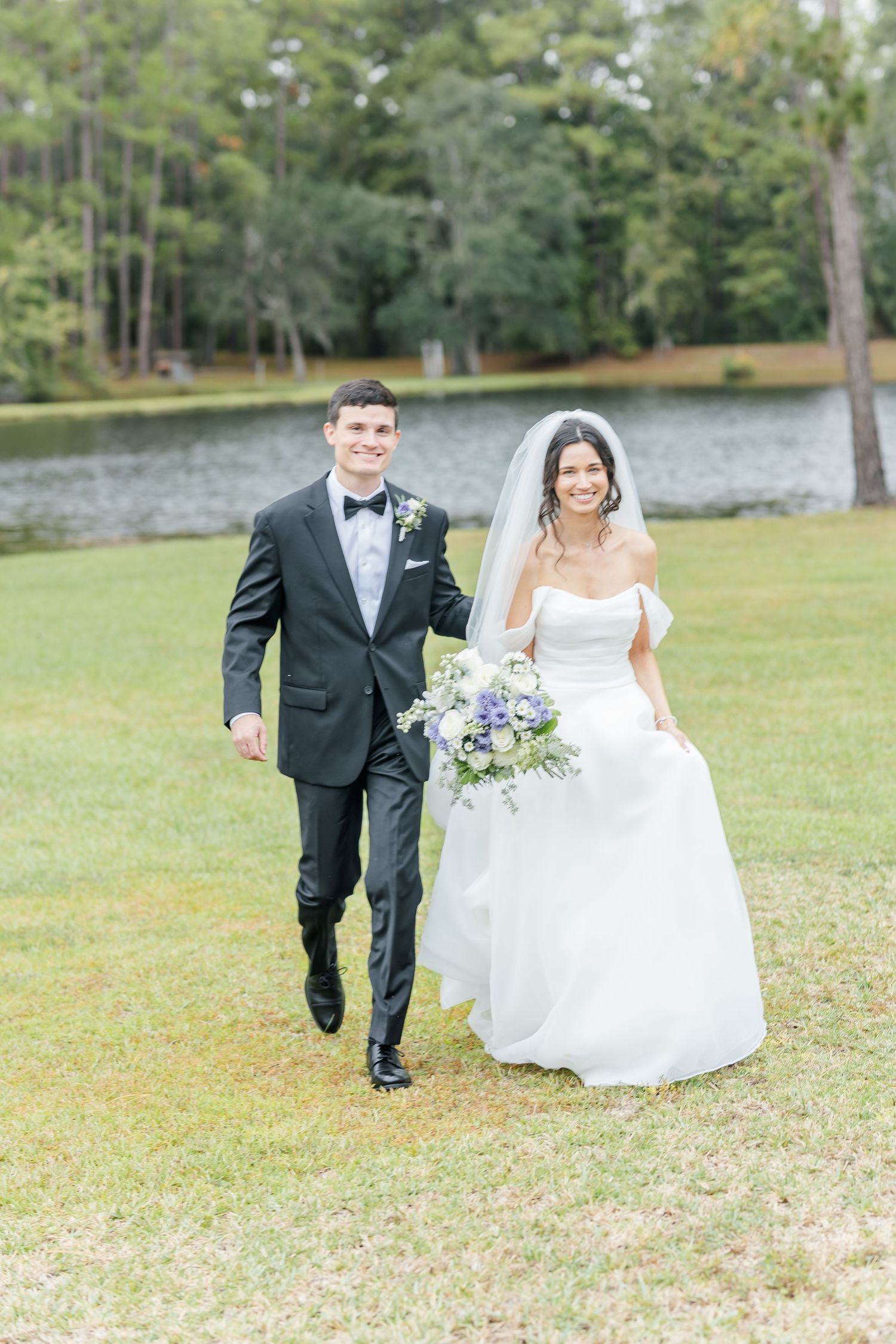 A couple in wedding attire playfully runs together across a grassy field with trees in the background.