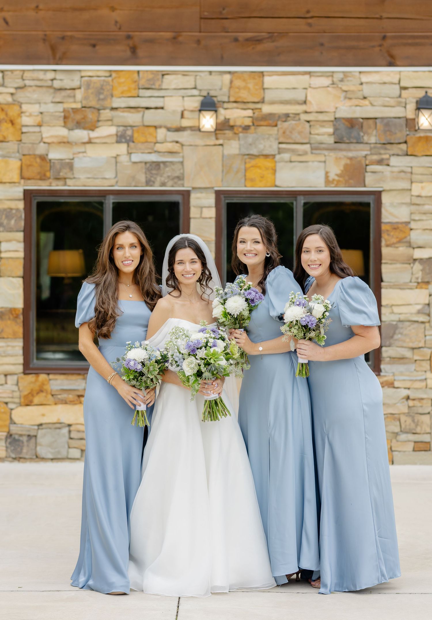 Wedding party in light blue bridesmaid dresses and white bridal gown pose in front of stone building.
