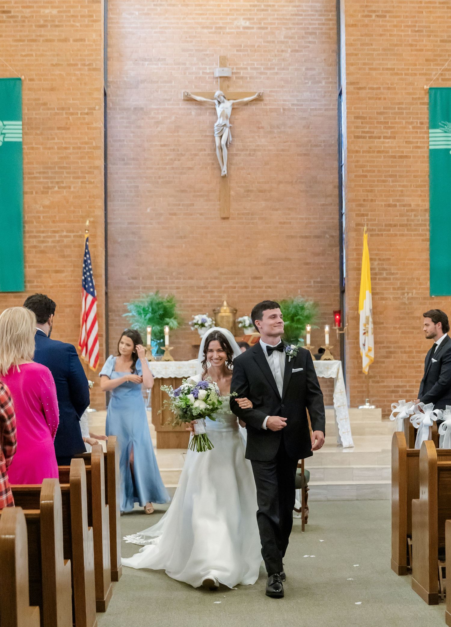 A traditional Catholic church wedding ceremony with guests seated in wooden pews and crucifixes on brick walls.
