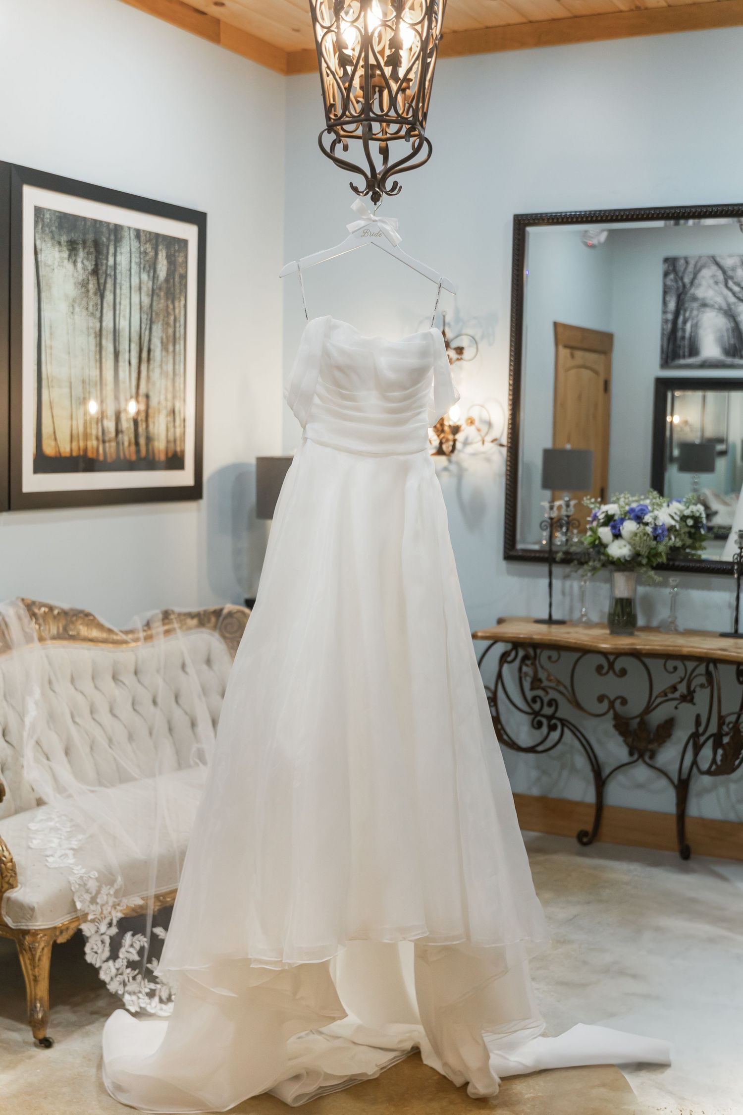 White wedding dress displayed in an elegant bridal boutique with ornate chandeliers and tufted furniture.