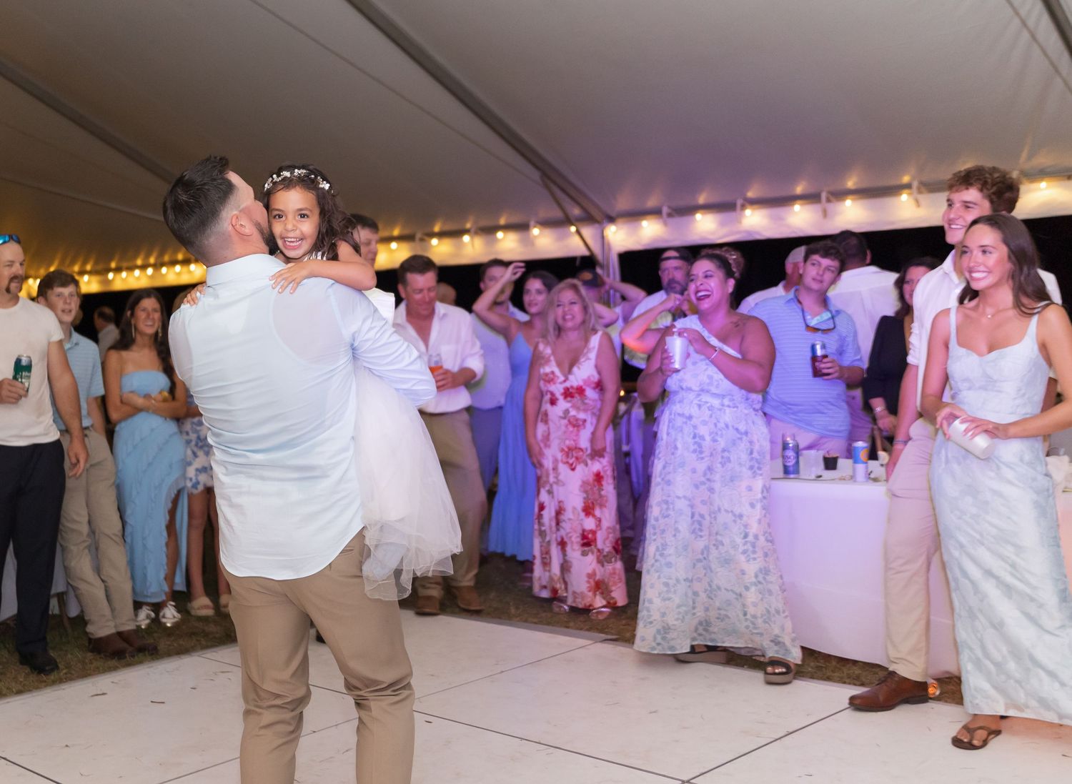 Wedding guests dancing in a line at reception under tent with purple lighting.