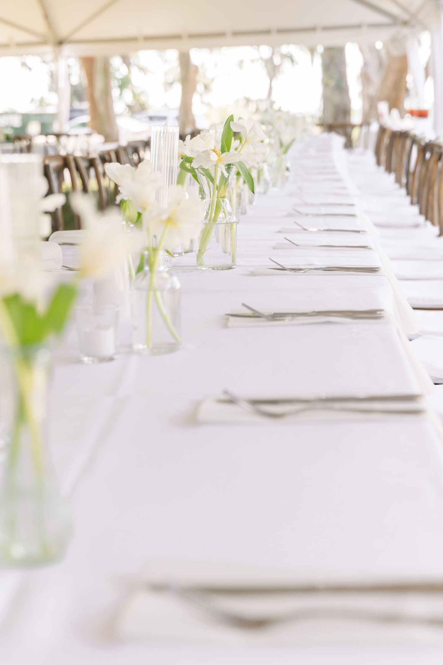 White tablescape with minimalist place settings and simple white flower arrangements in glass vases under a tent.