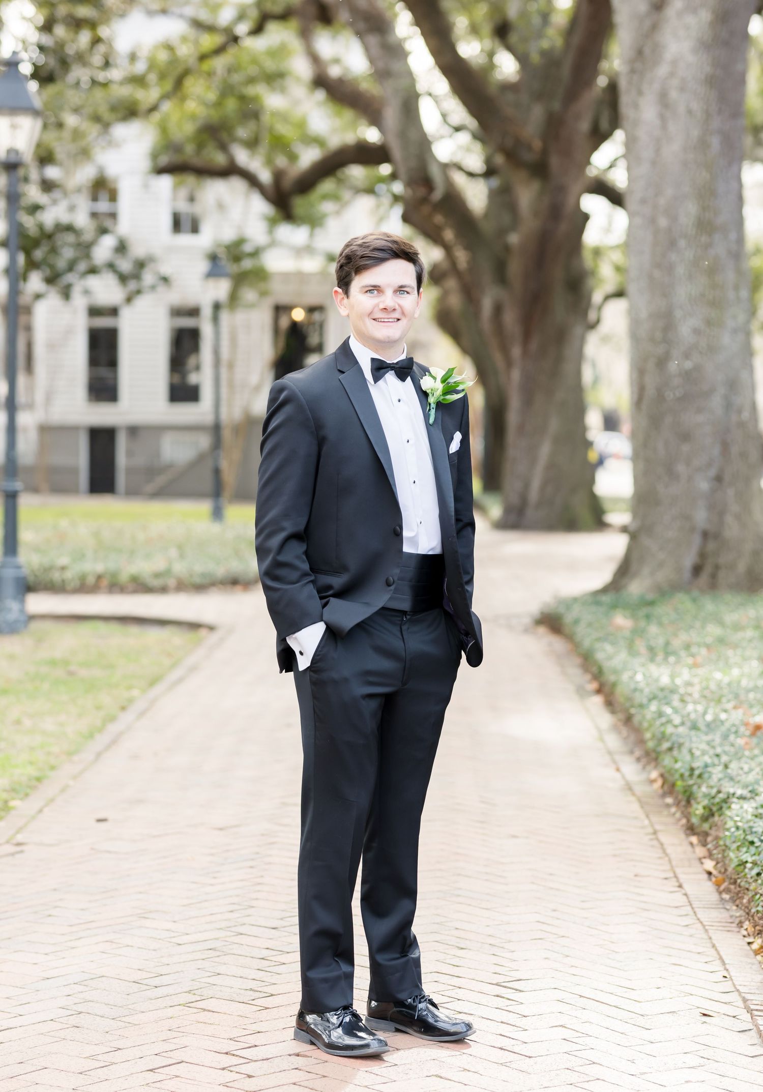 Groomsmen in black tuxedos pose casually on brick pathway for wedding photos.