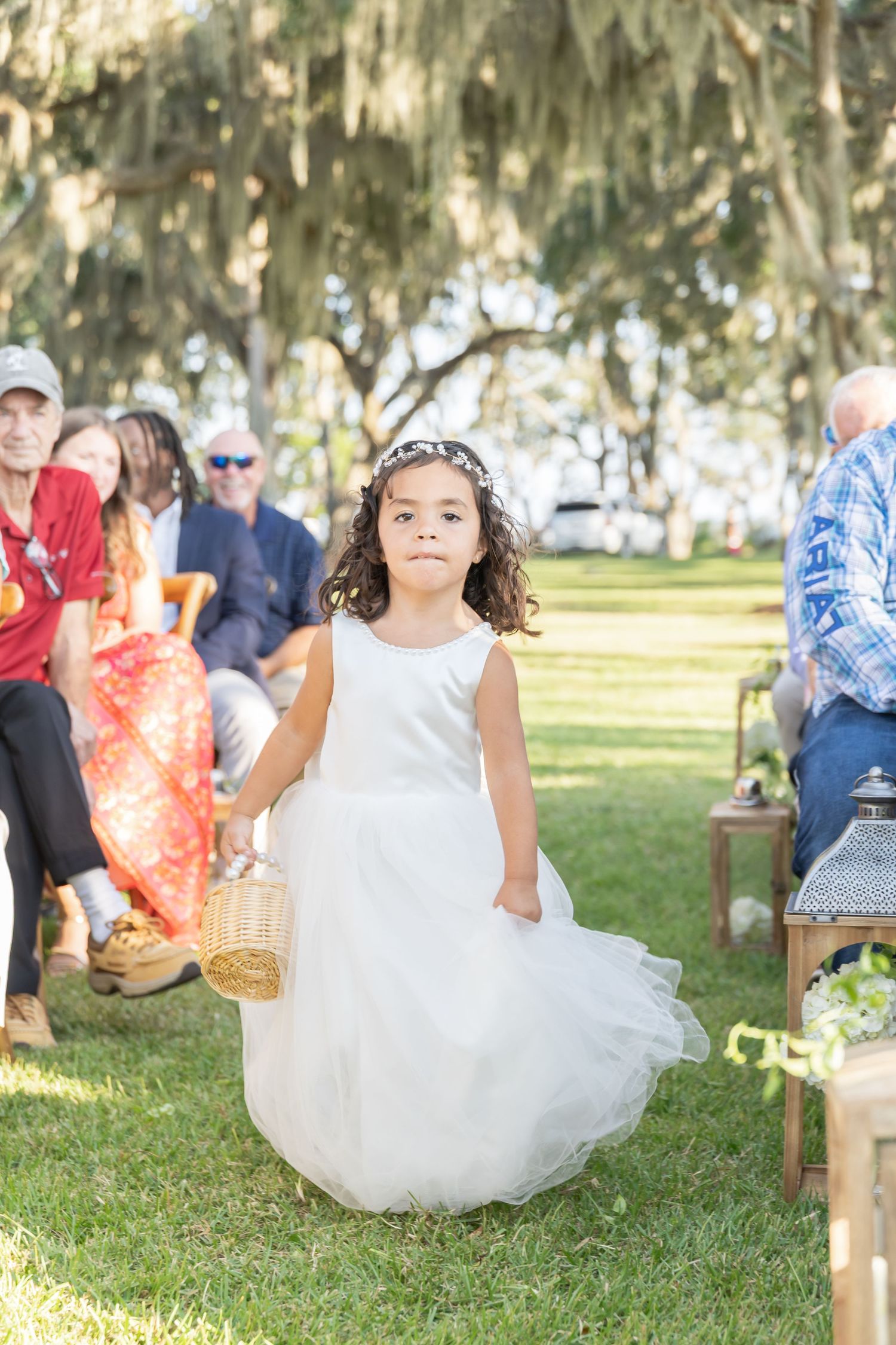 A small child in a white tulle dress walks down an outdoor aisle during a ceremony under Spanish moss draped trees.