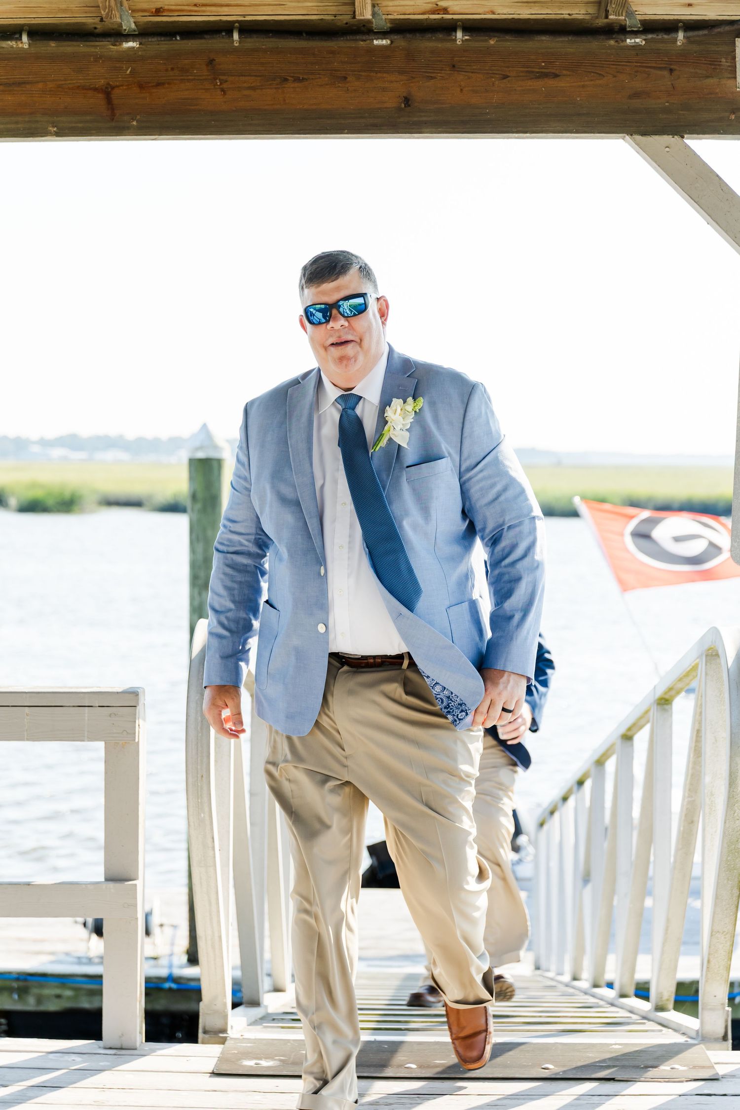 A groomsman in a light blue blazer and khakis strikes playful poses on a wooden dock by the water.