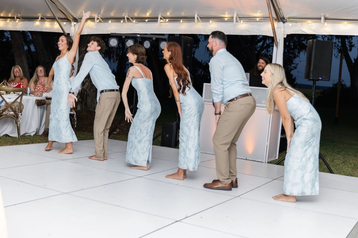 Wedding guests dance and celebrate under a white tent during an outdoor evening reception.