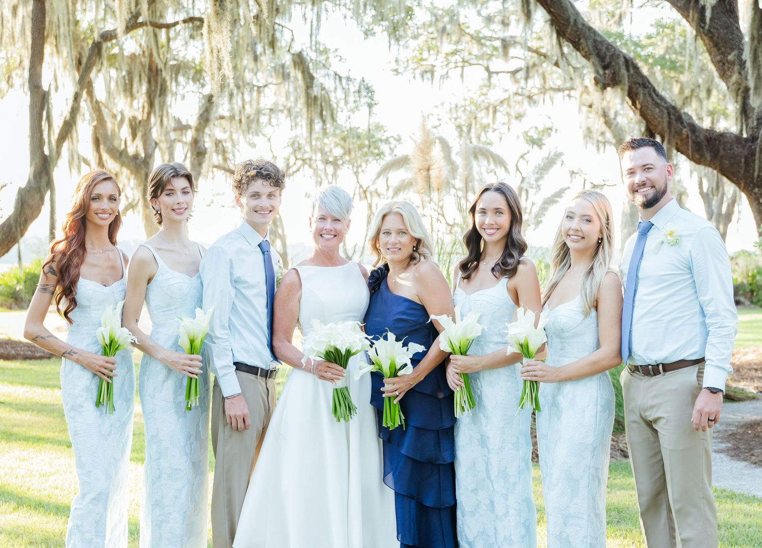 A wedding party poses under Spanish moss draped trees wearing white and blue attire for outdoor photos.