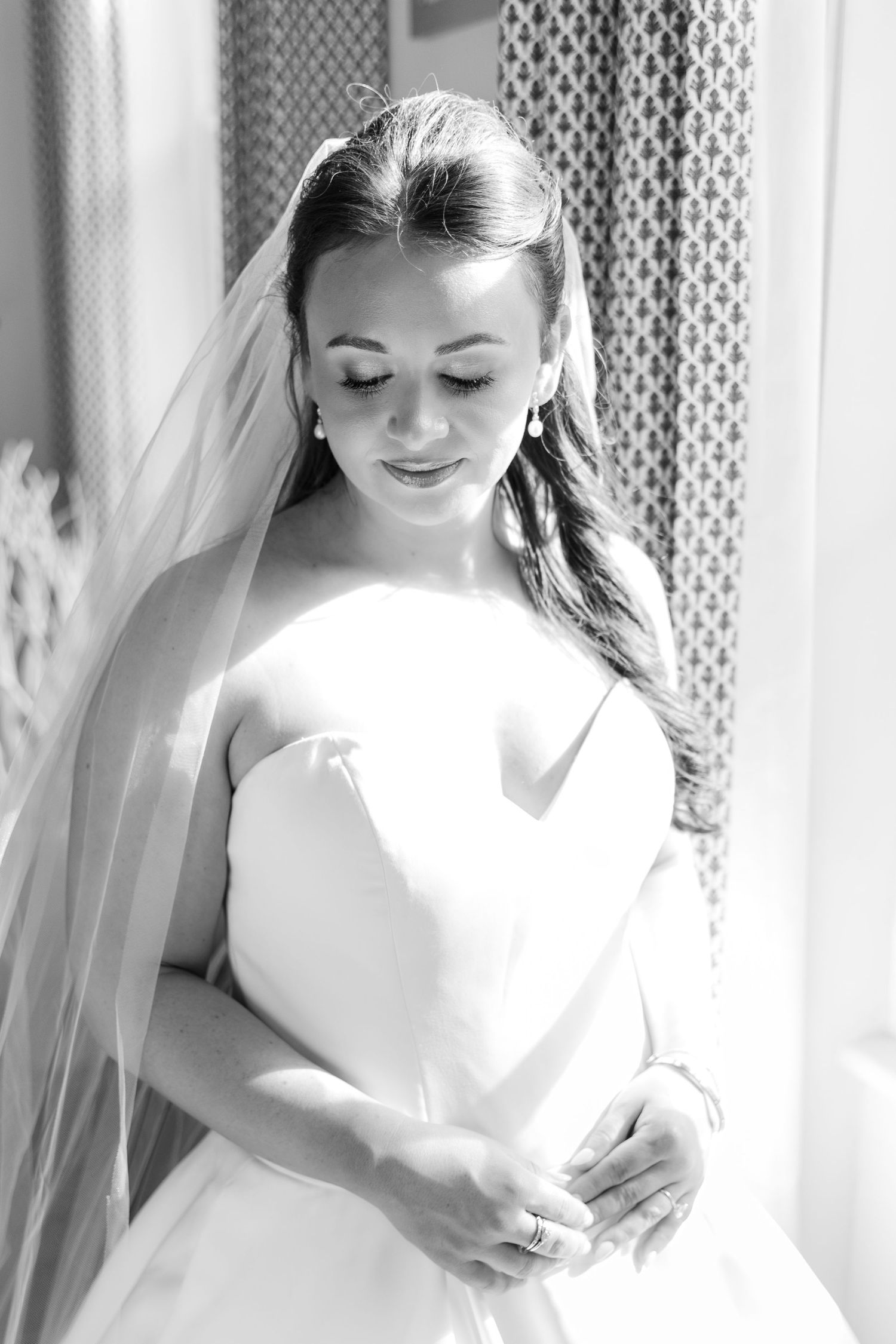 A bride in a white wedding dress smiles softly while looking down in an elegant black and white portrait.