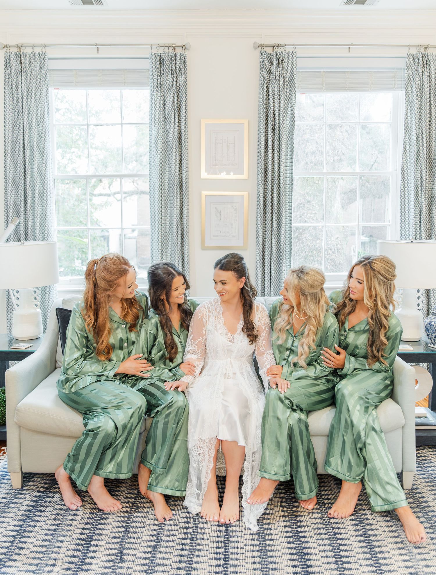 Wedding party wearing matching sage green robes lounge together on a white couch in a bright sunlit room.