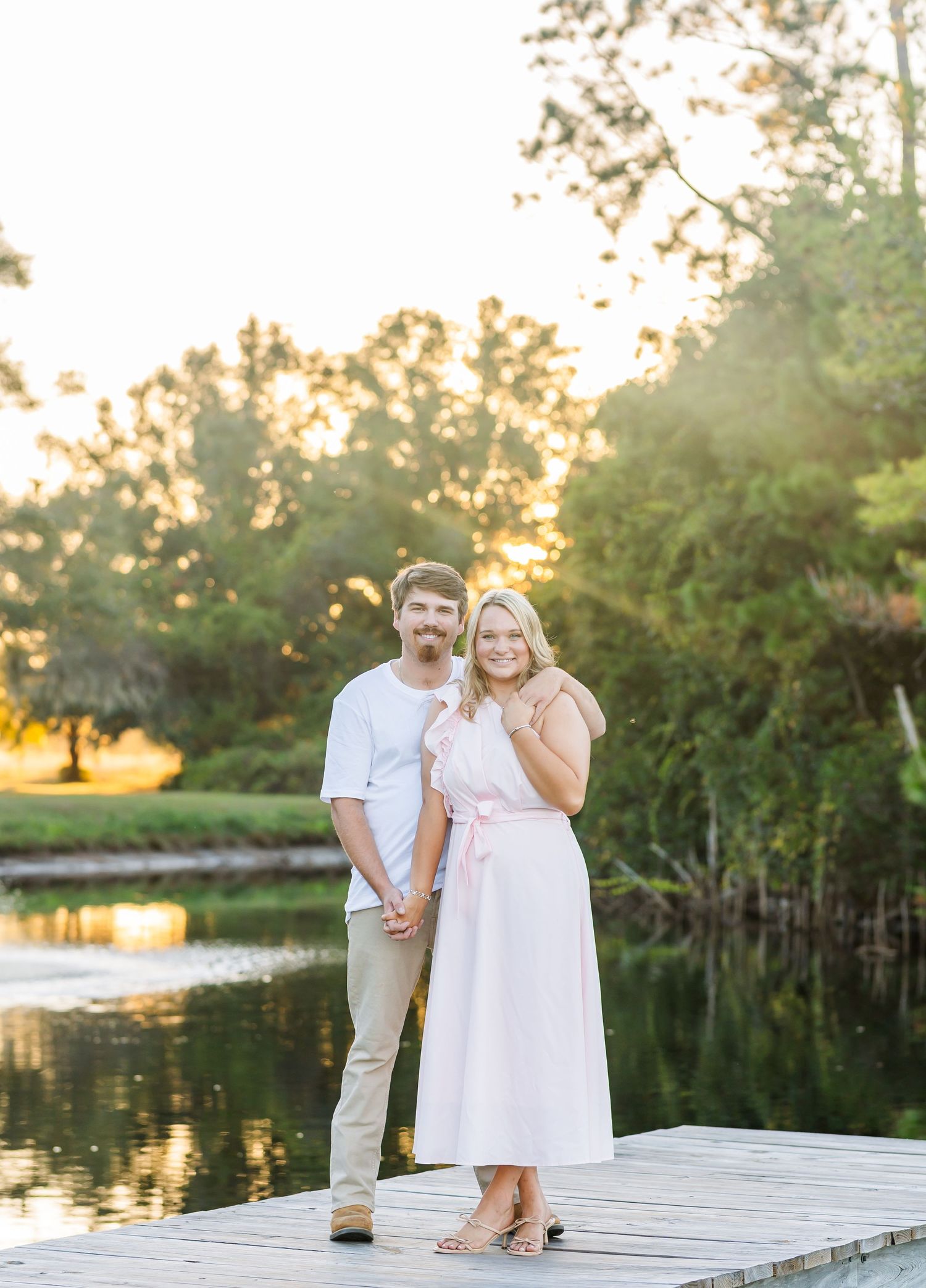 A couple poses together on a wooden dock beside a serene lake at golden hour.