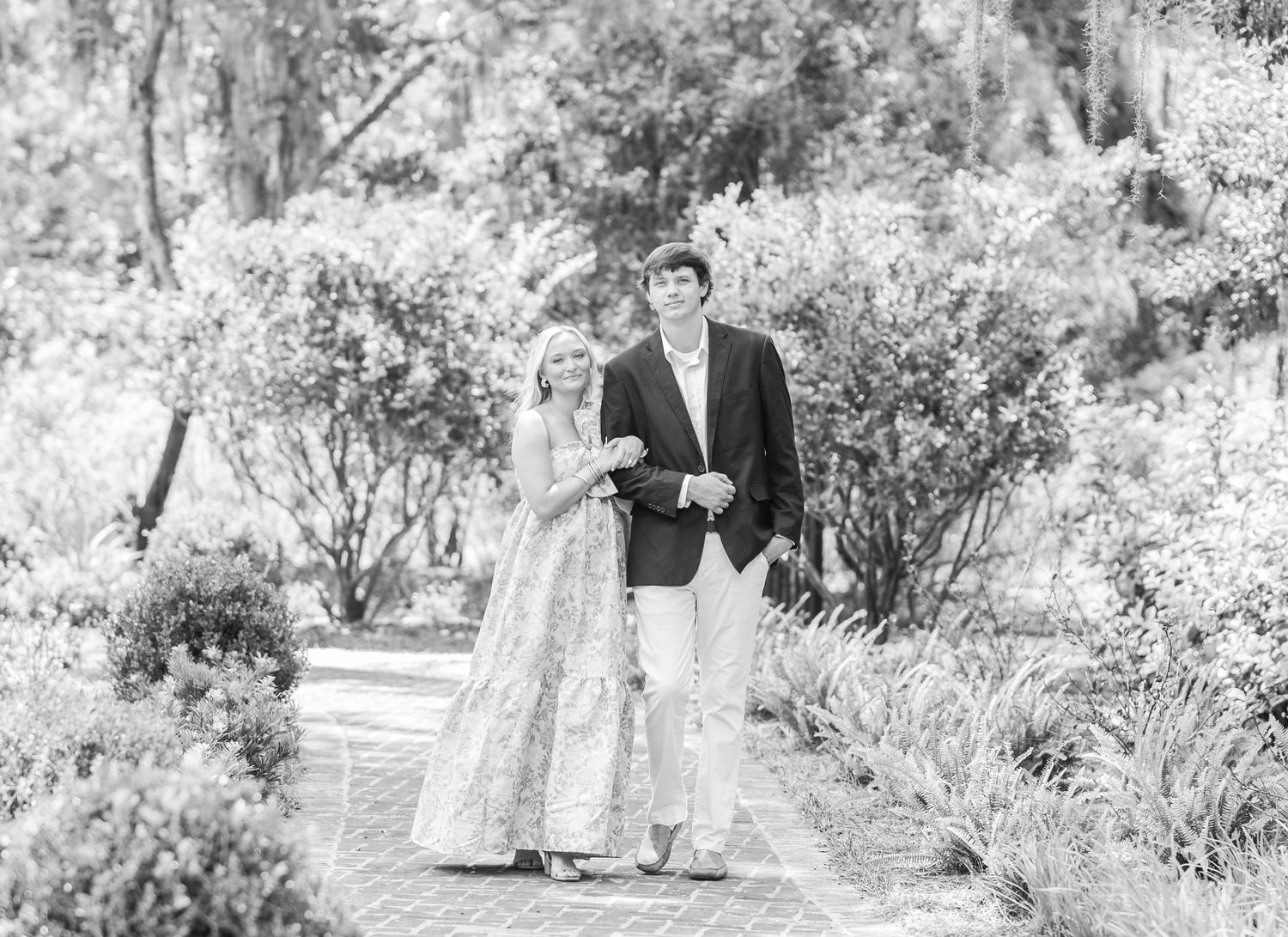 Black and white photo of a couple walking together in a garden setting with lush trees and foliage.