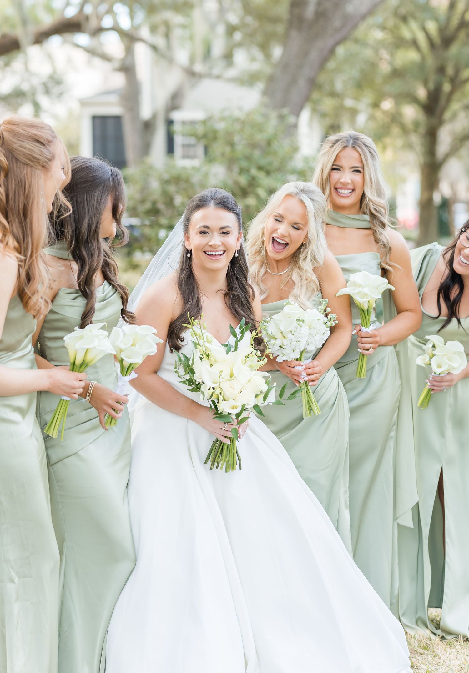 Bridesmaids in sage green dresses share joyful laughter with bride holding white bouquets in outdoor garden setting.