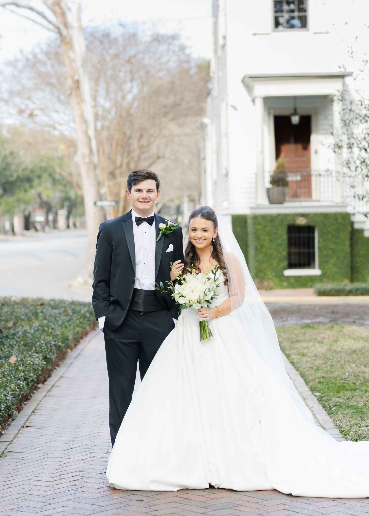 Elegant wedding couple poses together on a brick path surrounded by historic buildings and greenery.