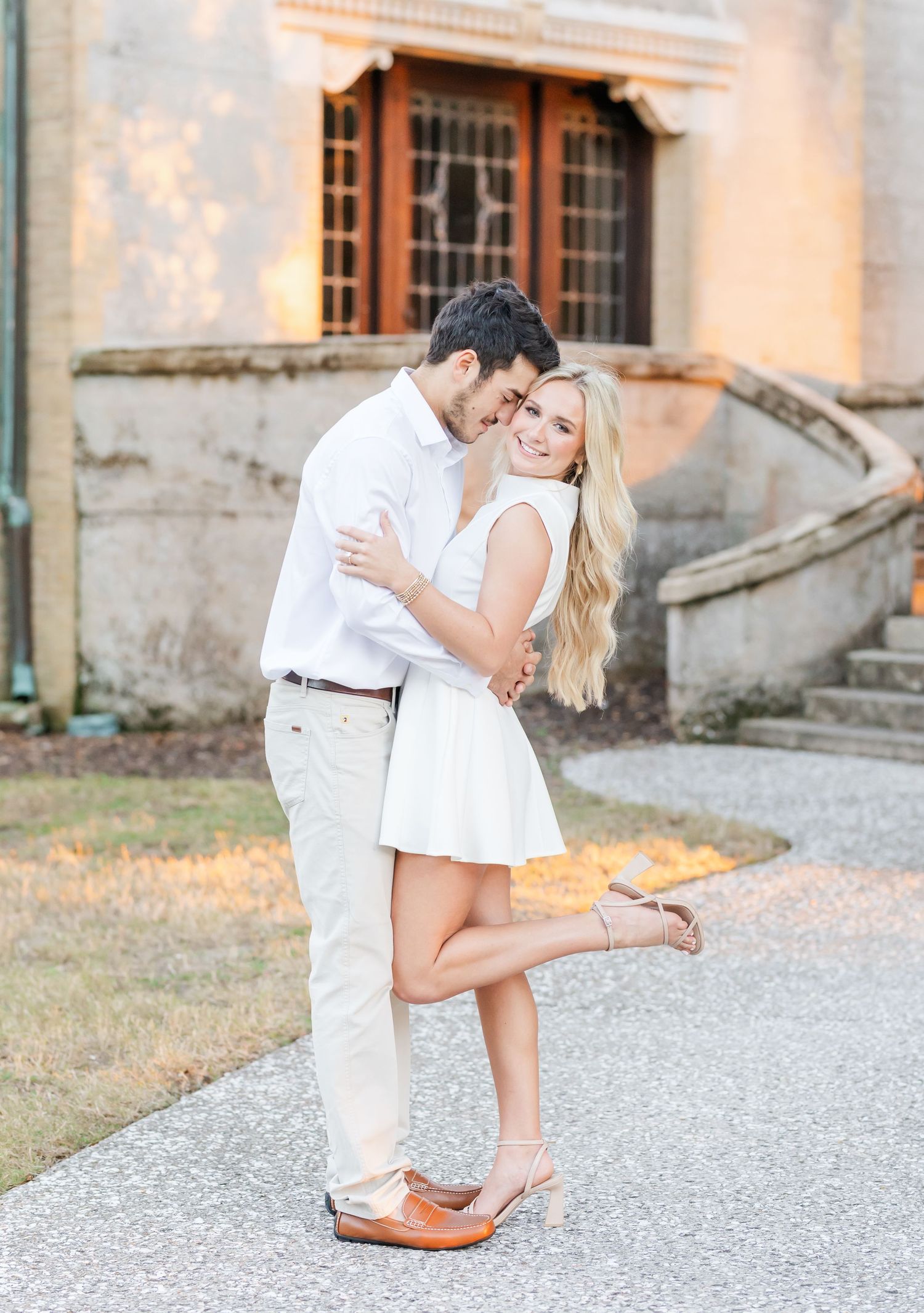 A romantic couple in white outfits shares intimate moments along a gravel pathway at dusk.