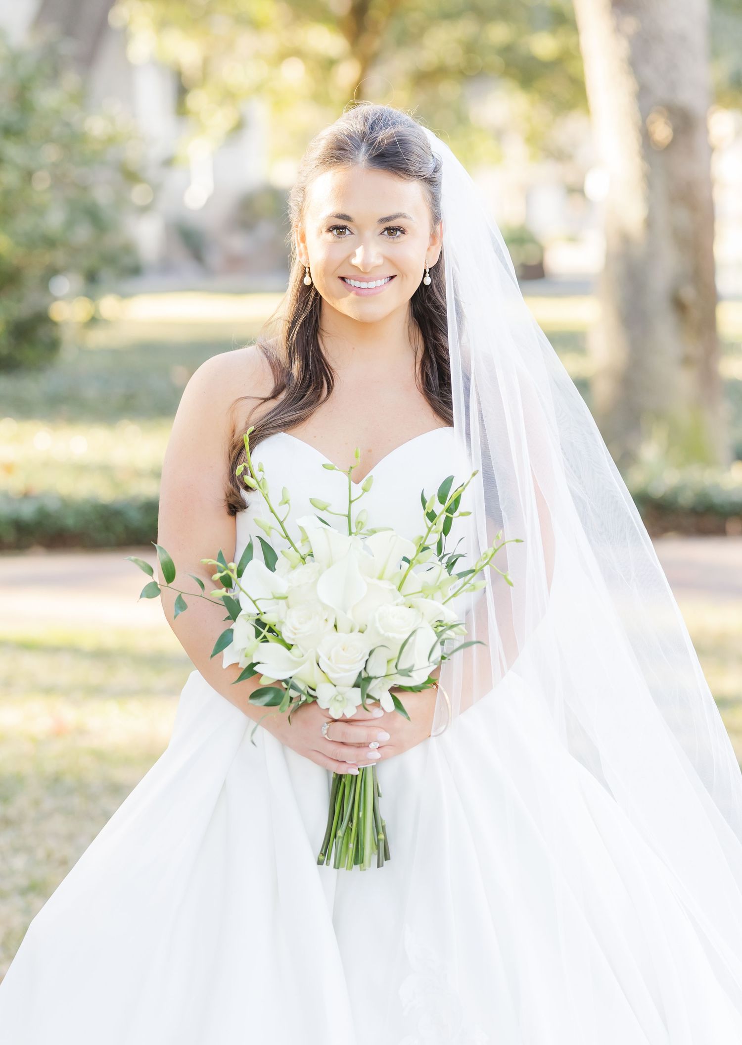 Elegant bride in white strapless gown holds classic white lily bouquet during outdoor wedding photoshoot.