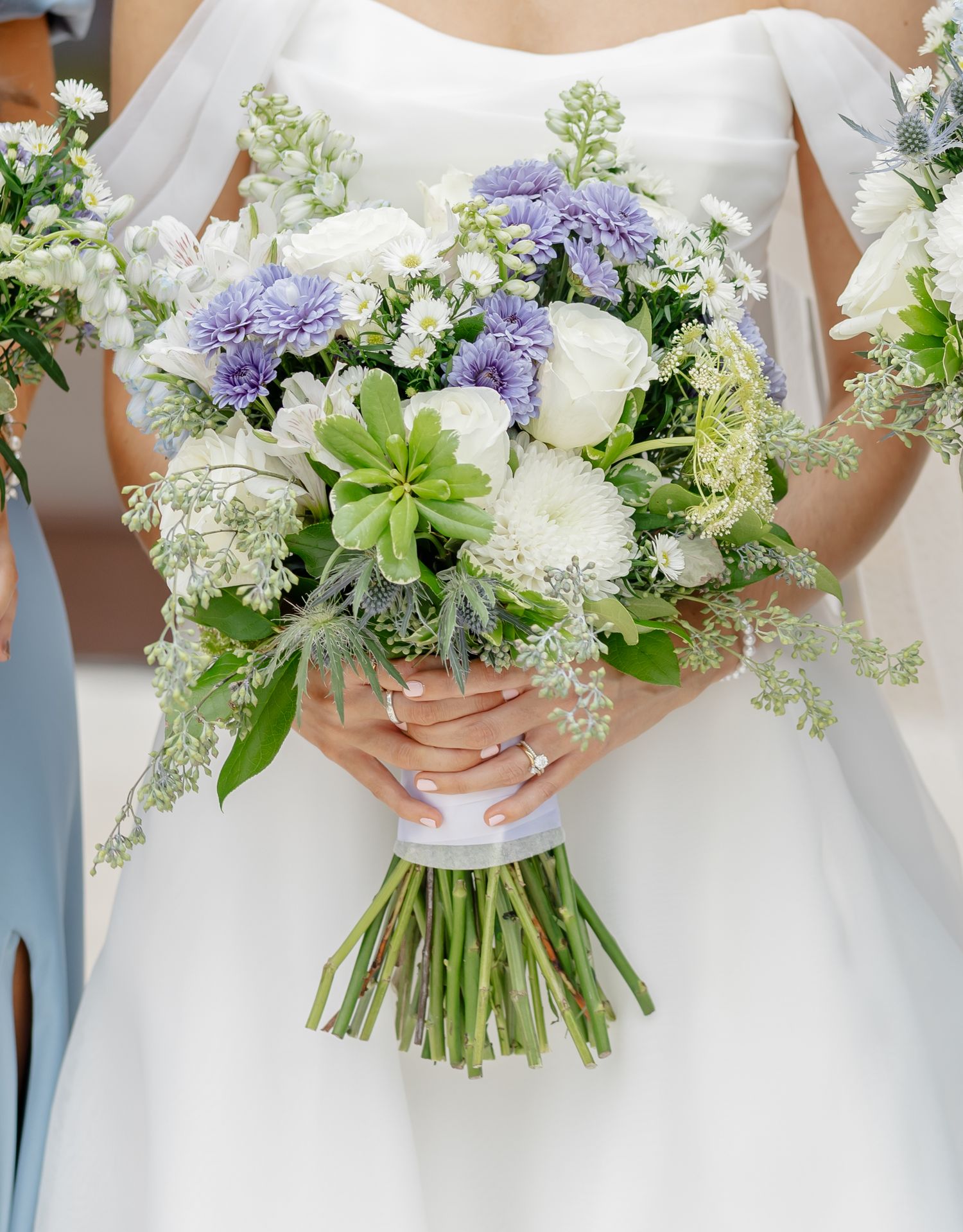 A bridal bouquet featuring white roses, purple hydrangeas, succulents and delicate greenery tied with a white ribbon.