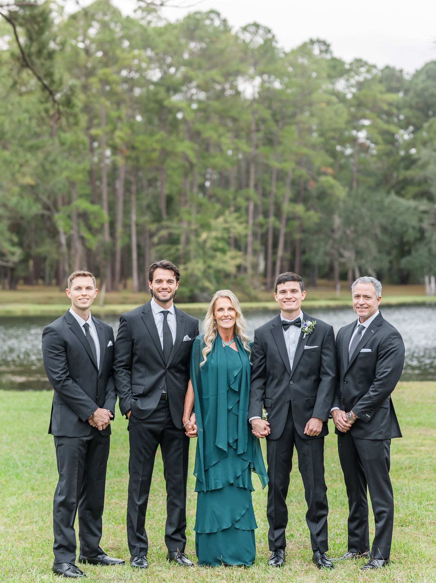 Wedding party members in black suits and emerald green dress pose together on grassy lawn with pine trees in background.