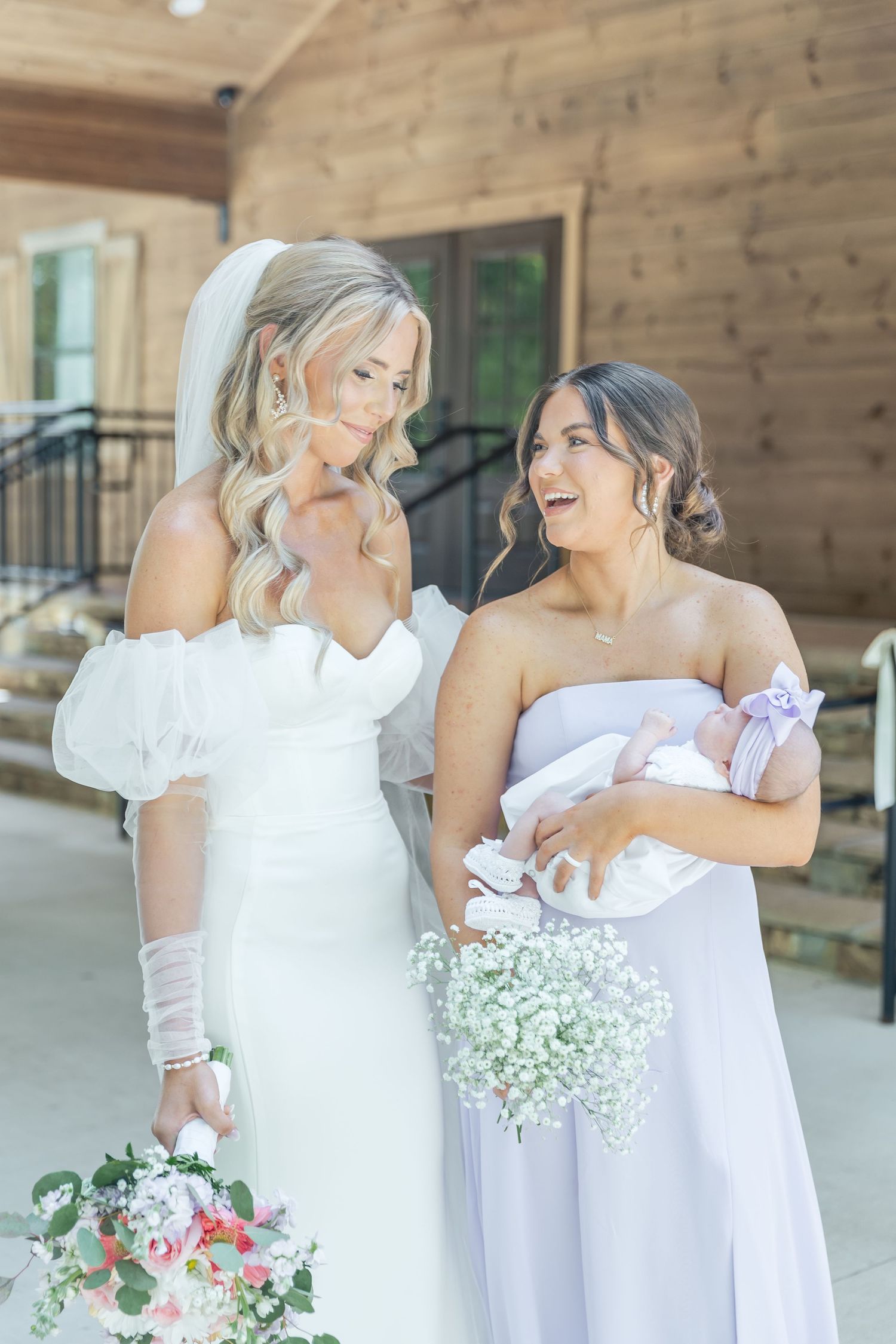 Two women in bridal attire share joyful moment while holding baby and bouquet.
