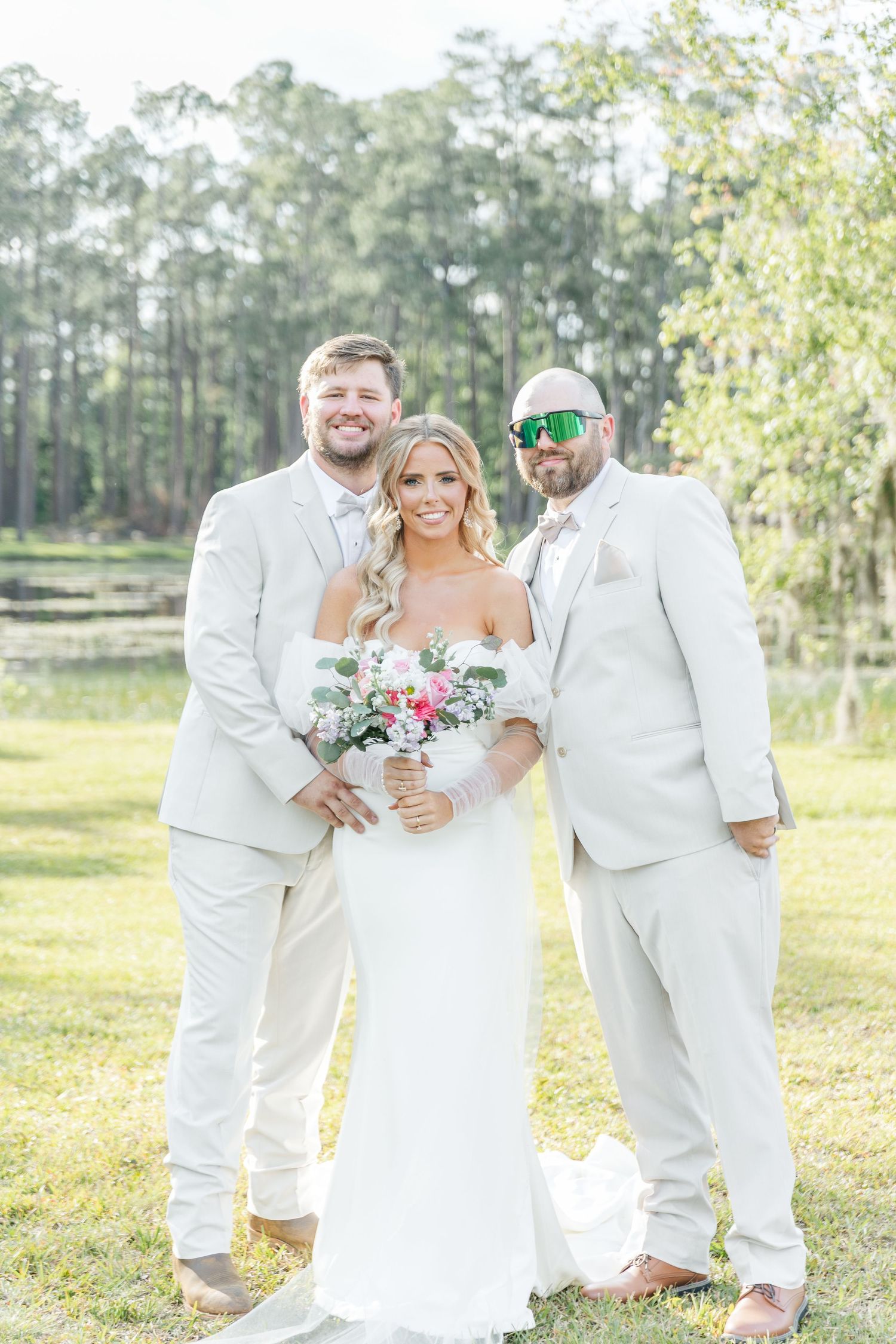 Wedding party poses outdoors in all-white attire with pink and white bouquet against natural forest backdrop.