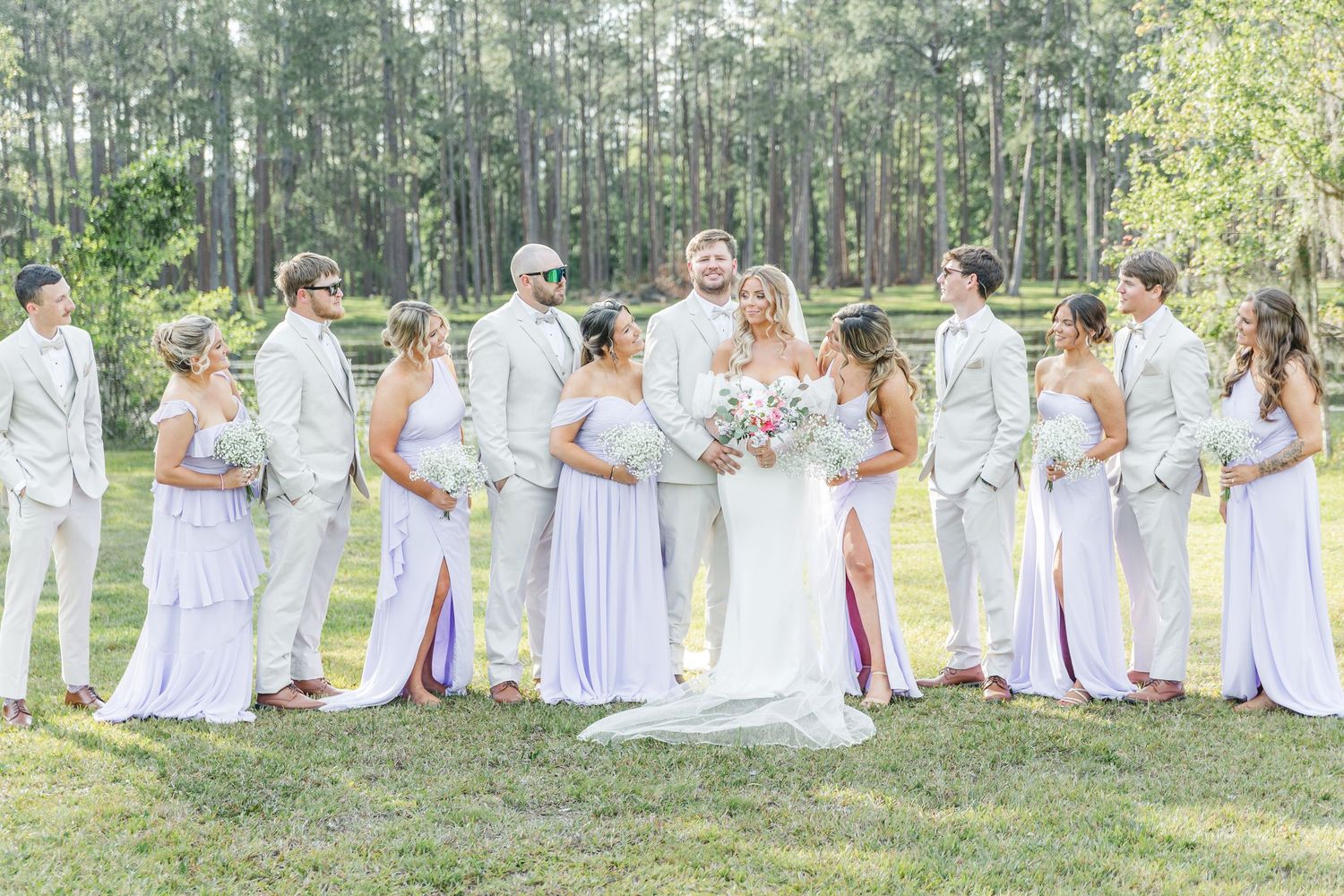 Bridal party in coordinating white and lavender outfits posed in a forest clearing.
