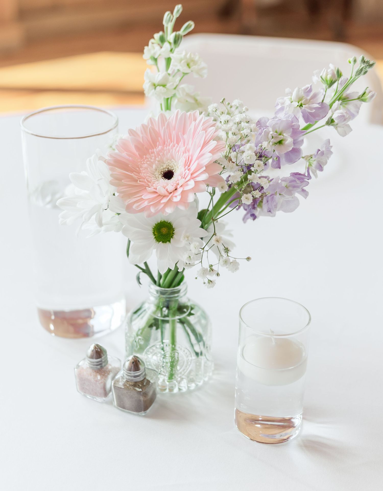 Simple glass vase centerpiece with pink gerbera daisies, white baby's breath, and purple flowers on white tablecloth.