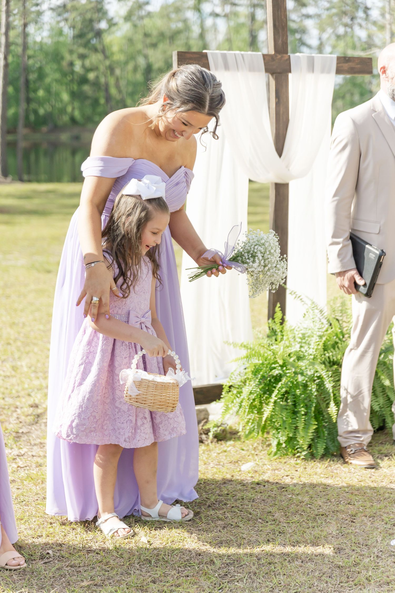 Bridesmaid in lavender dress helping flower girl with basket at outdoor ceremony.