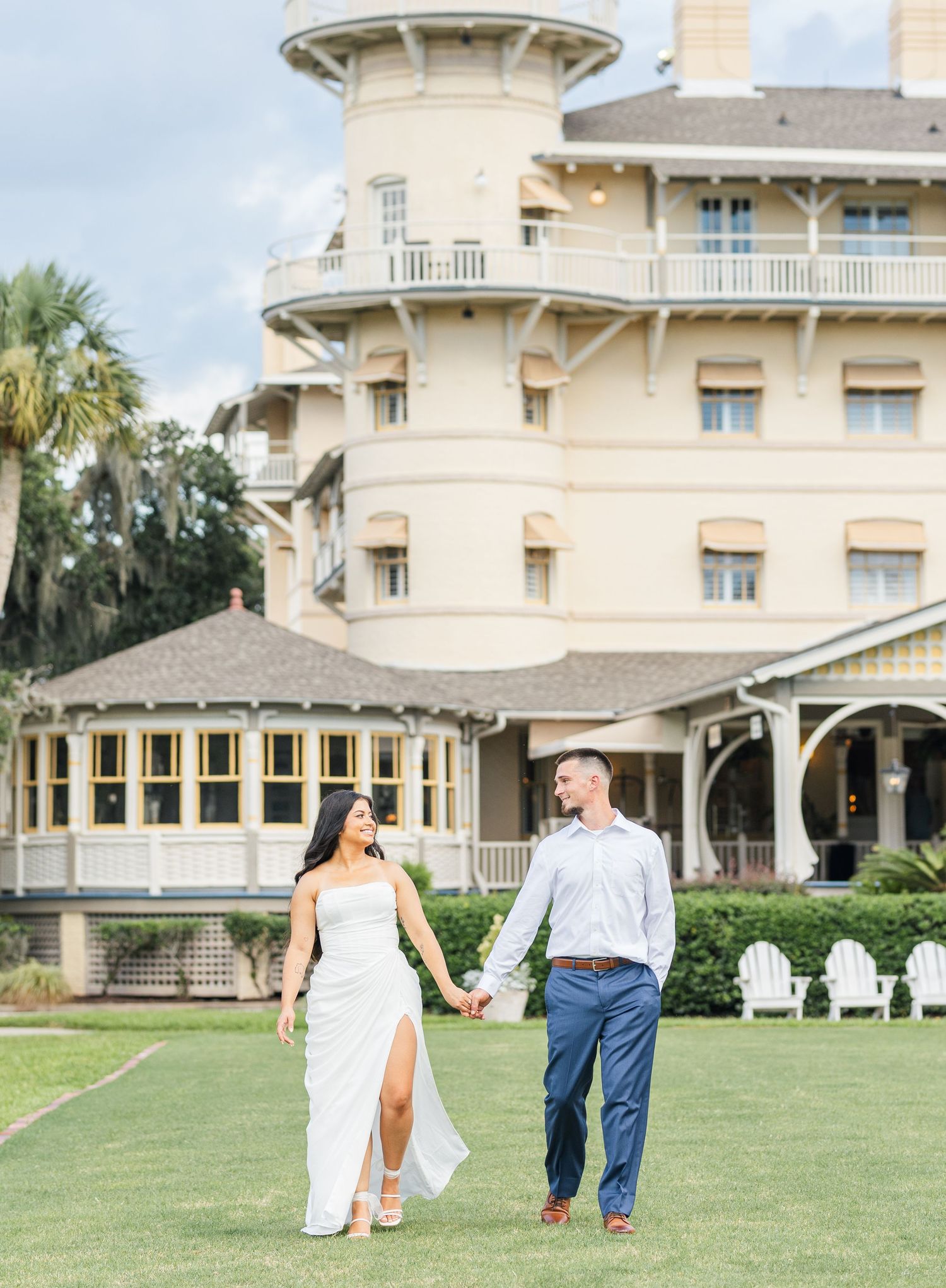 Elegant couple holding hands walking on lawn before a grand historic hotel building on a cloudy day.