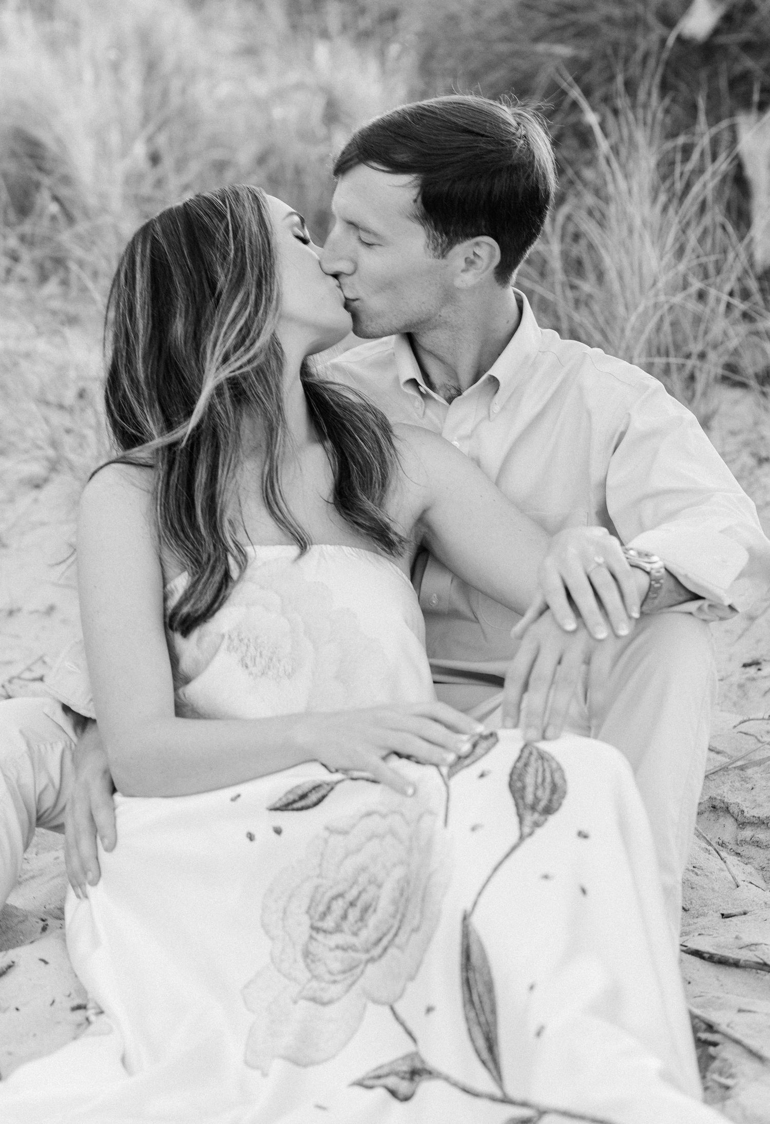 Black and white photo of a couple sharing a romantic kiss on a sandy beach, woman in floral dress.