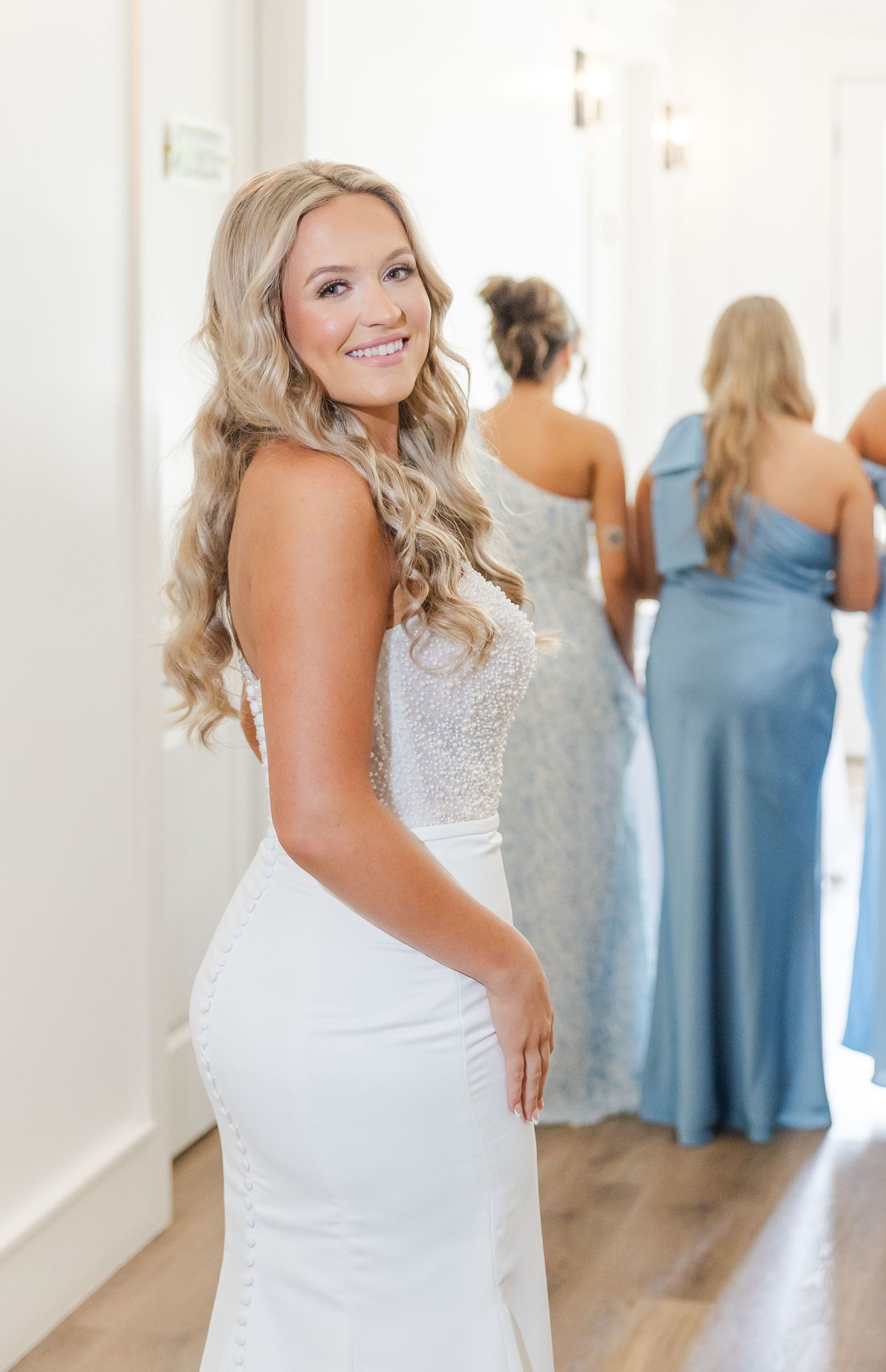 Bride in white beaded gown smiles over shoulder with bridesmaids in blue dresses visible in background.