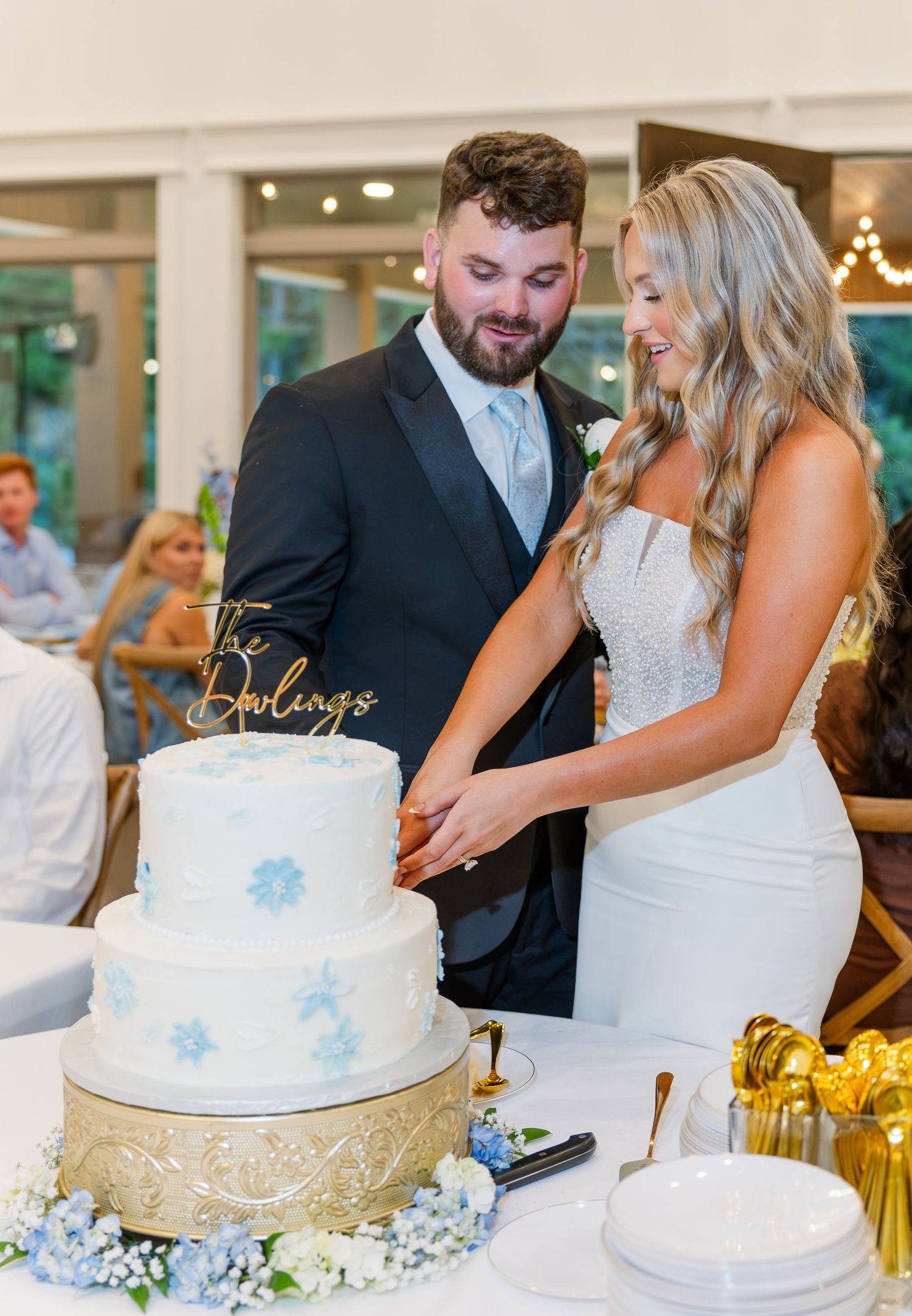 Bride and groom cut a two-tier white wedding cake decorated with blue flowers at reception.