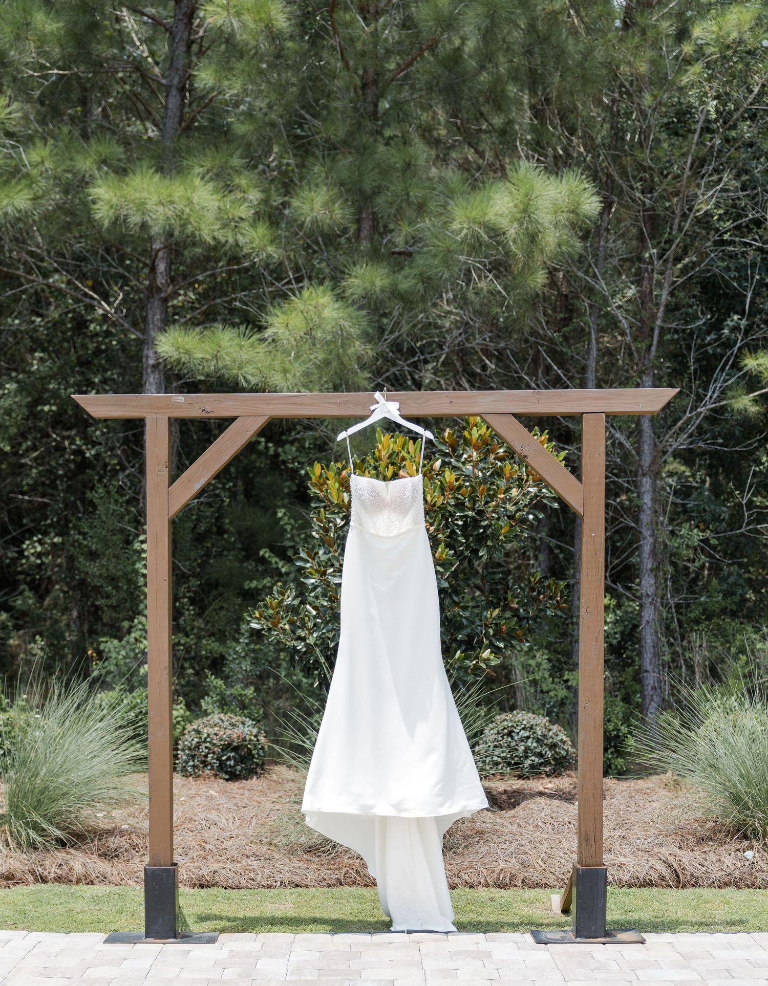 White strapless wedding gown hanging from a rustic wooden outdoor arch surrounded by pine trees.