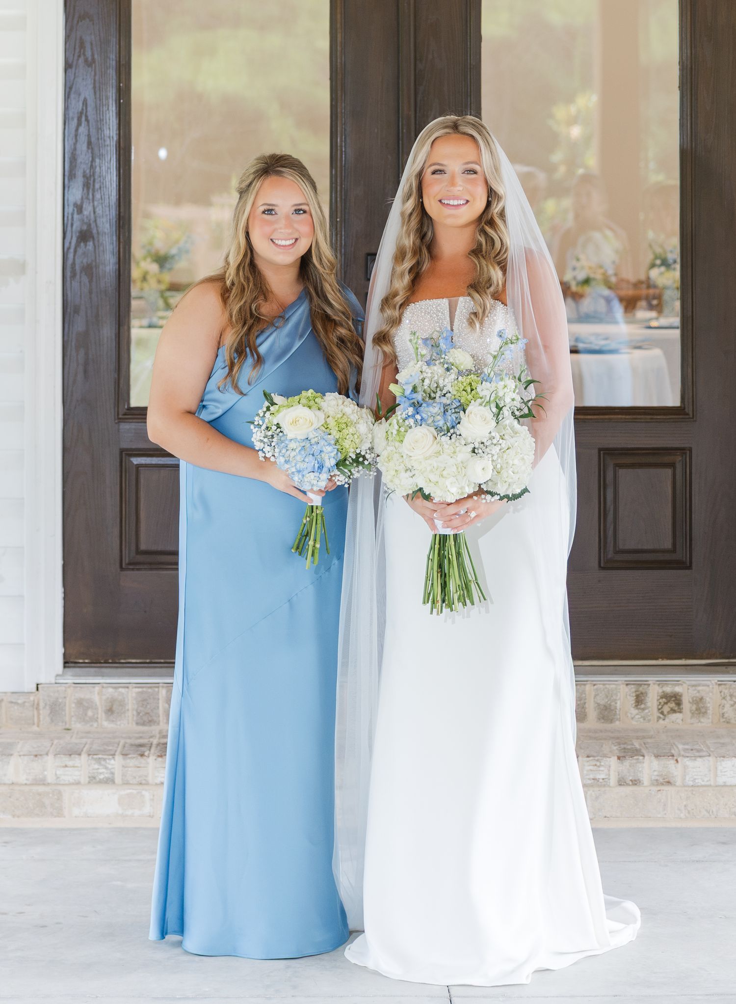 Bride in white lace gown and bridesmaid in blue dress holding hydrangea bouquets at wedding venue.